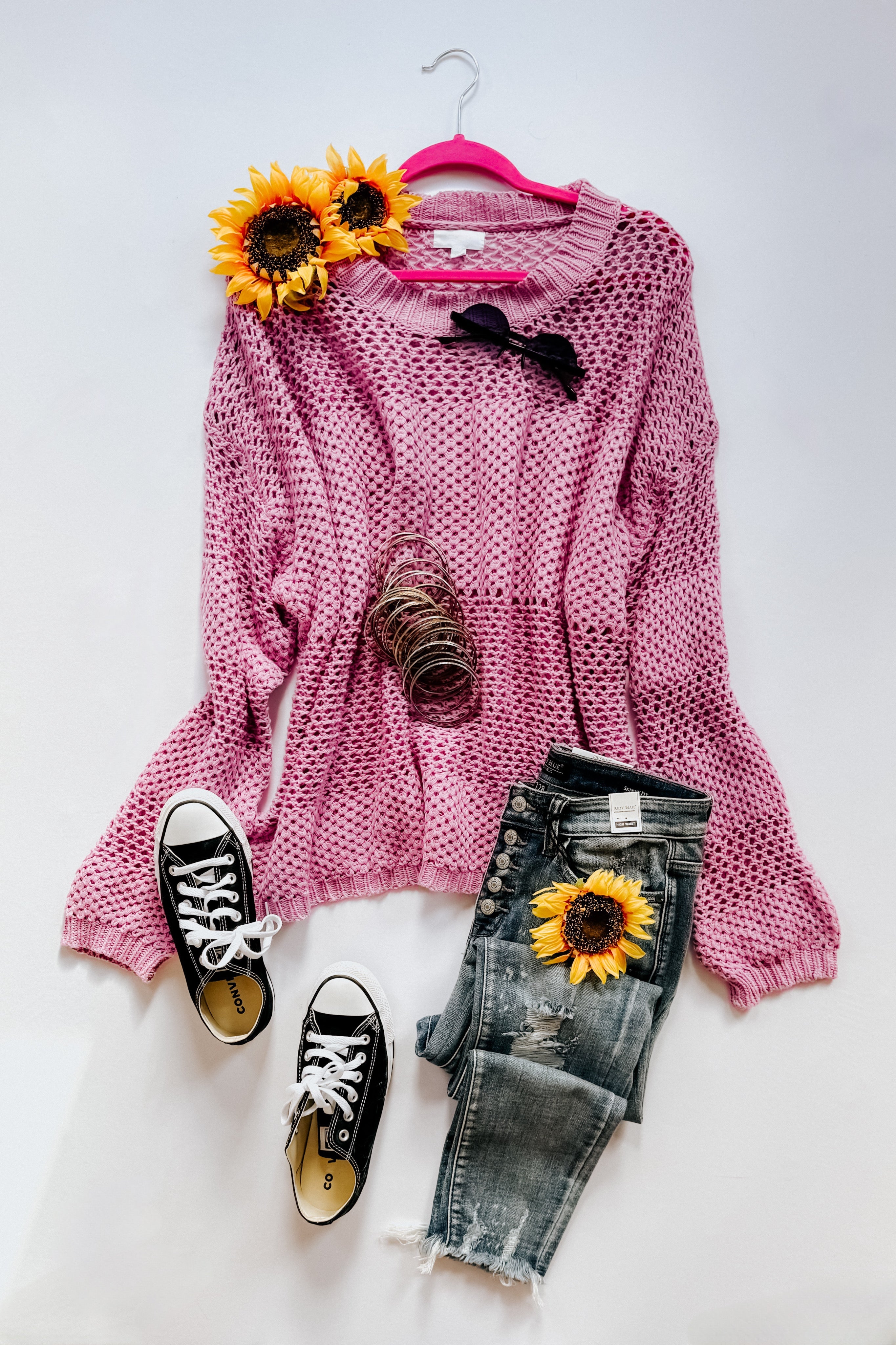Pink loose-knit sweater styled with distressed jeans, black sneakers, sunflower accents, and sunglasses on white background