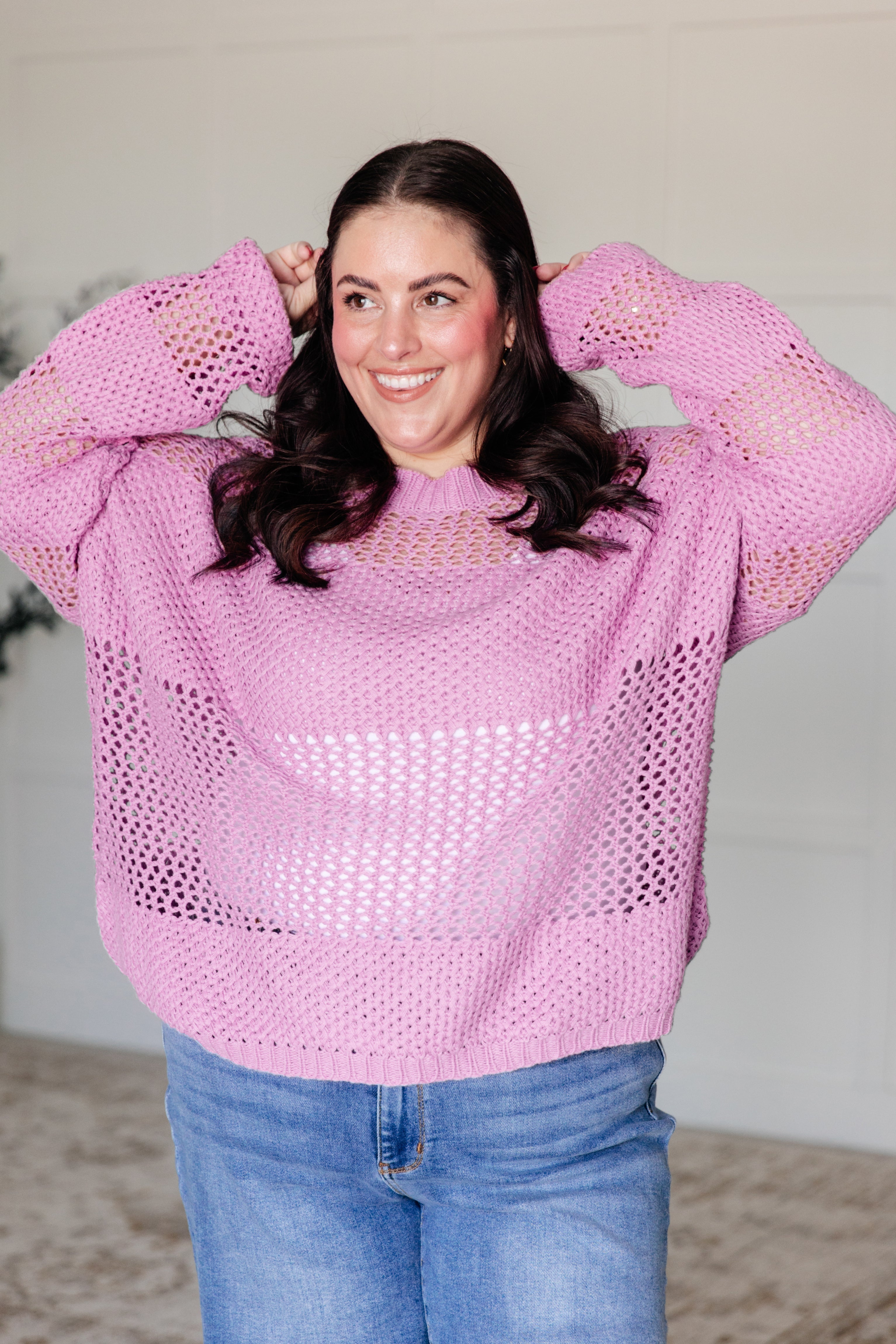 Plus-size woman smiling wearing a loose pink open knit sweater with light blue jeans indoors