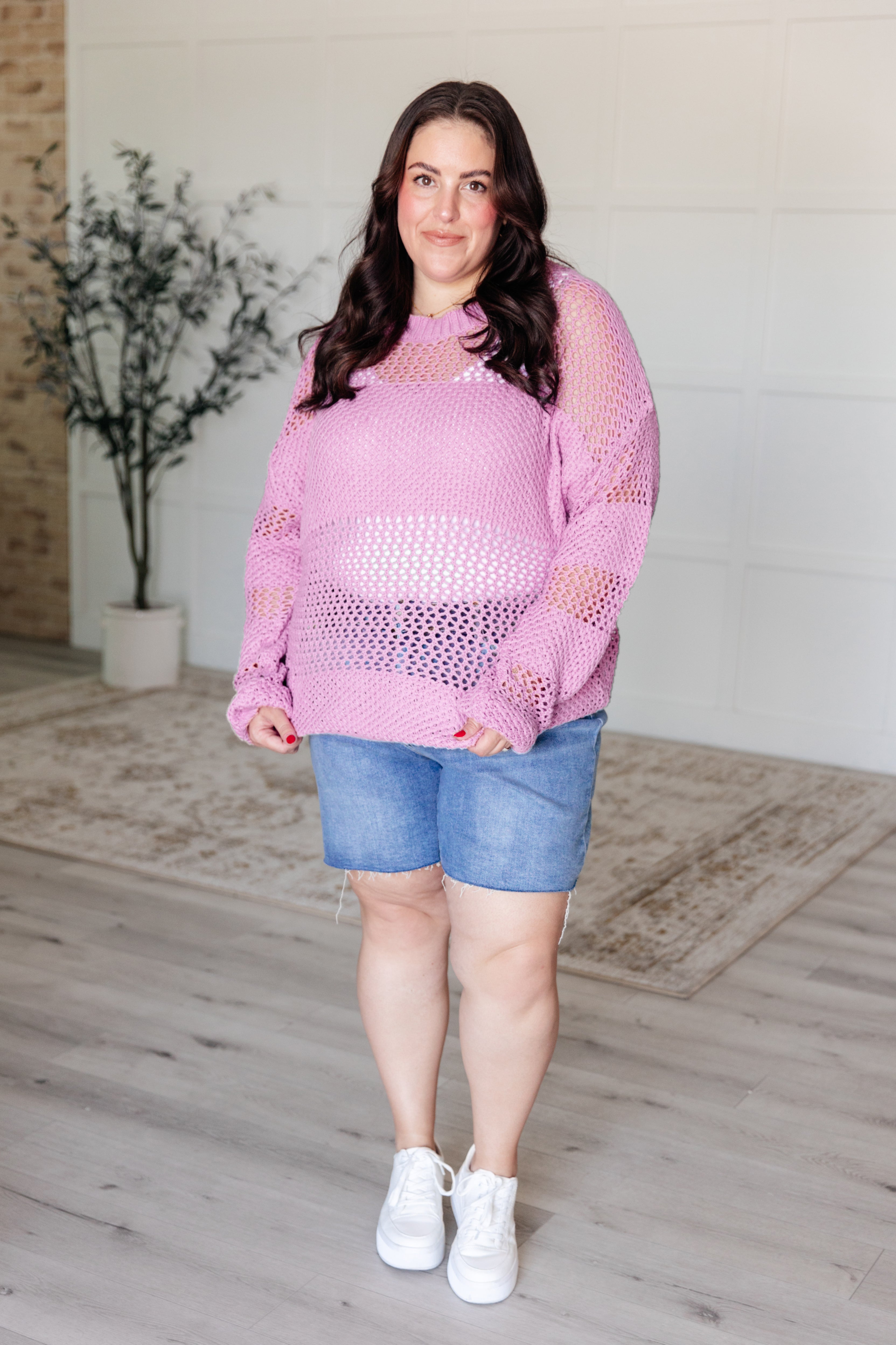 Plus-size woman wearing a loose pink open knit sweater paired with denim shorts and white sneakers indoors