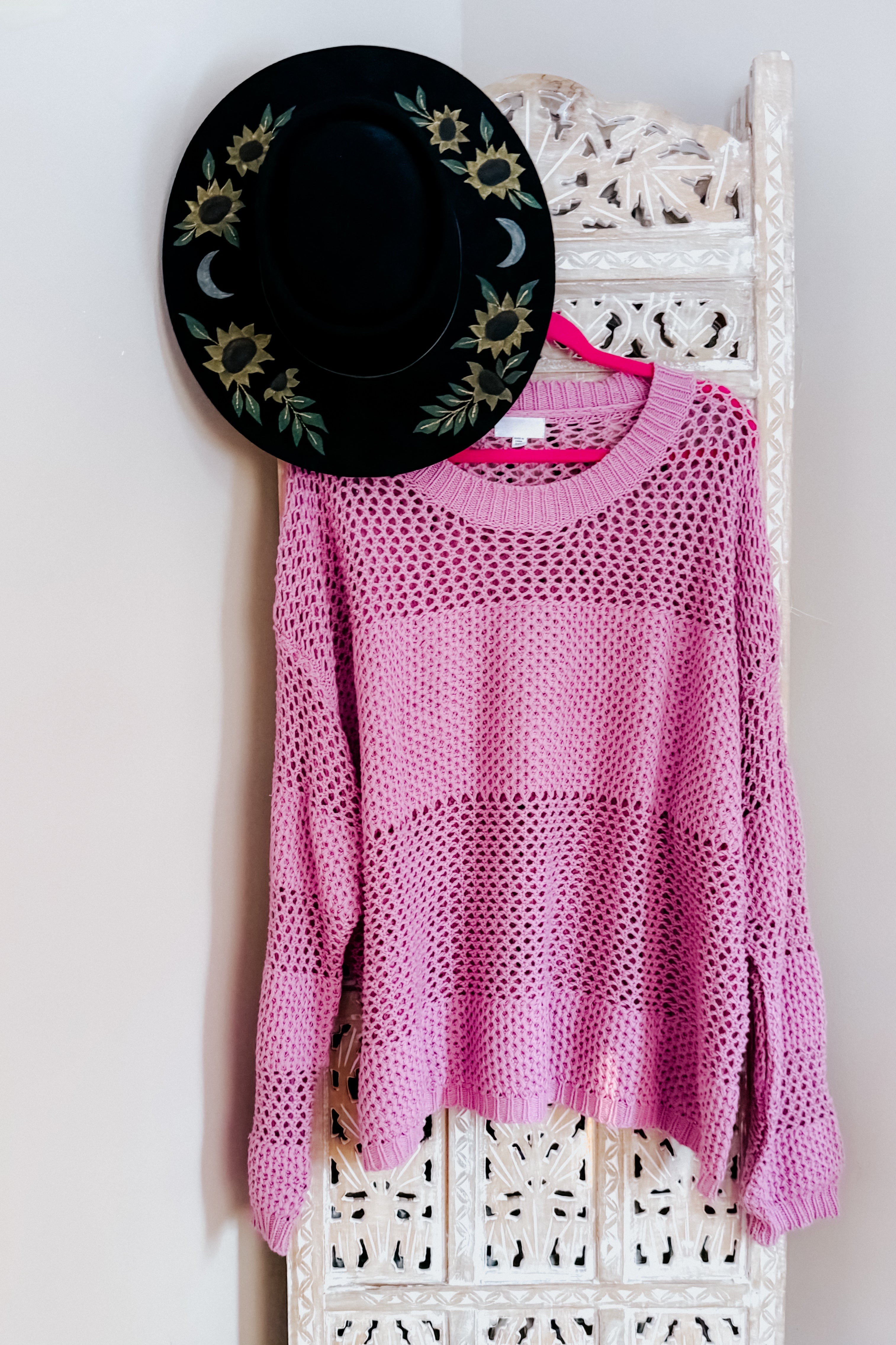Loose pink open-knit sweater displayed on a carved white wooden screen with a black floral hat hanging above