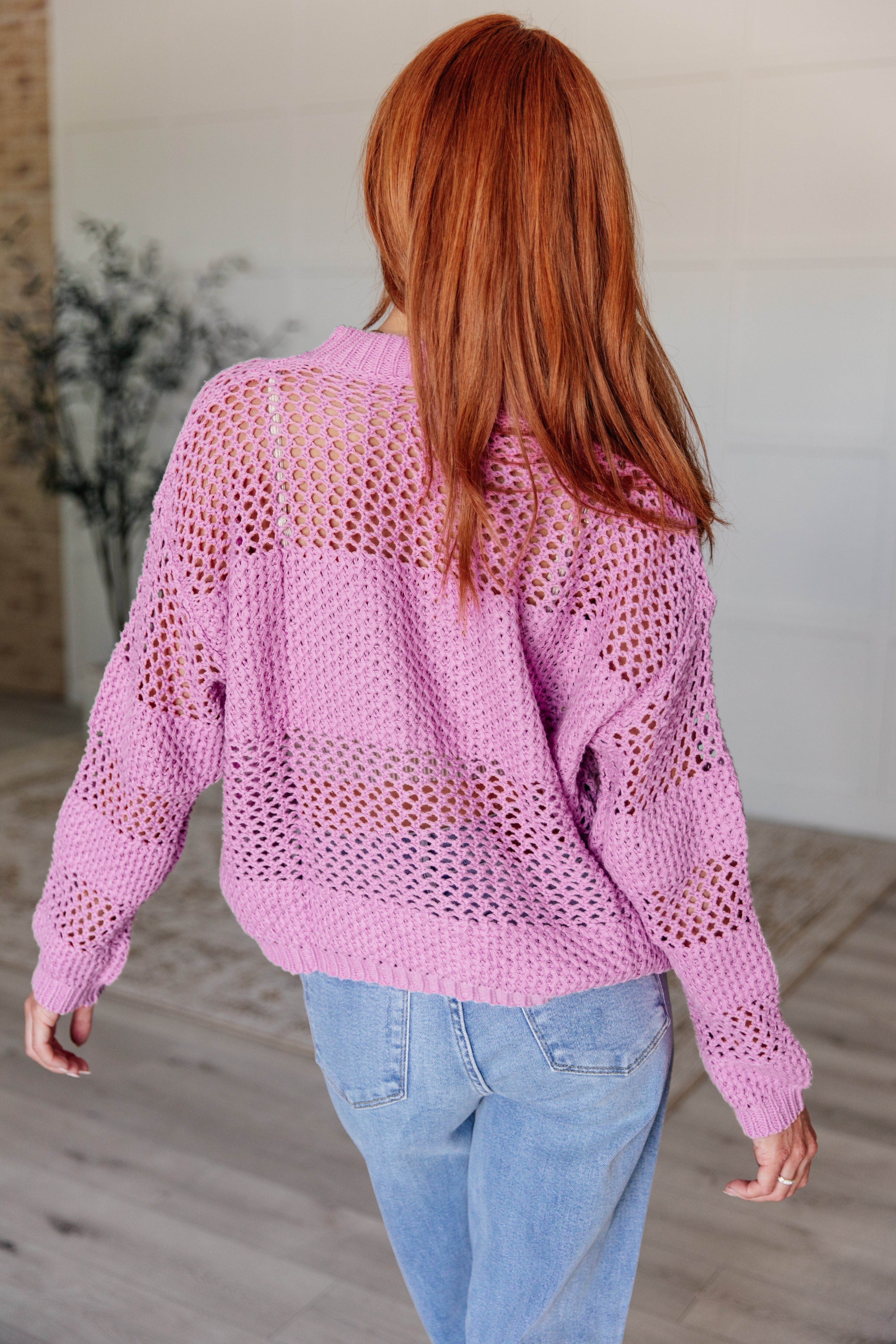 Woman with red hair wearing a loose lavender open knit sweater paired with light blue jeans indoors