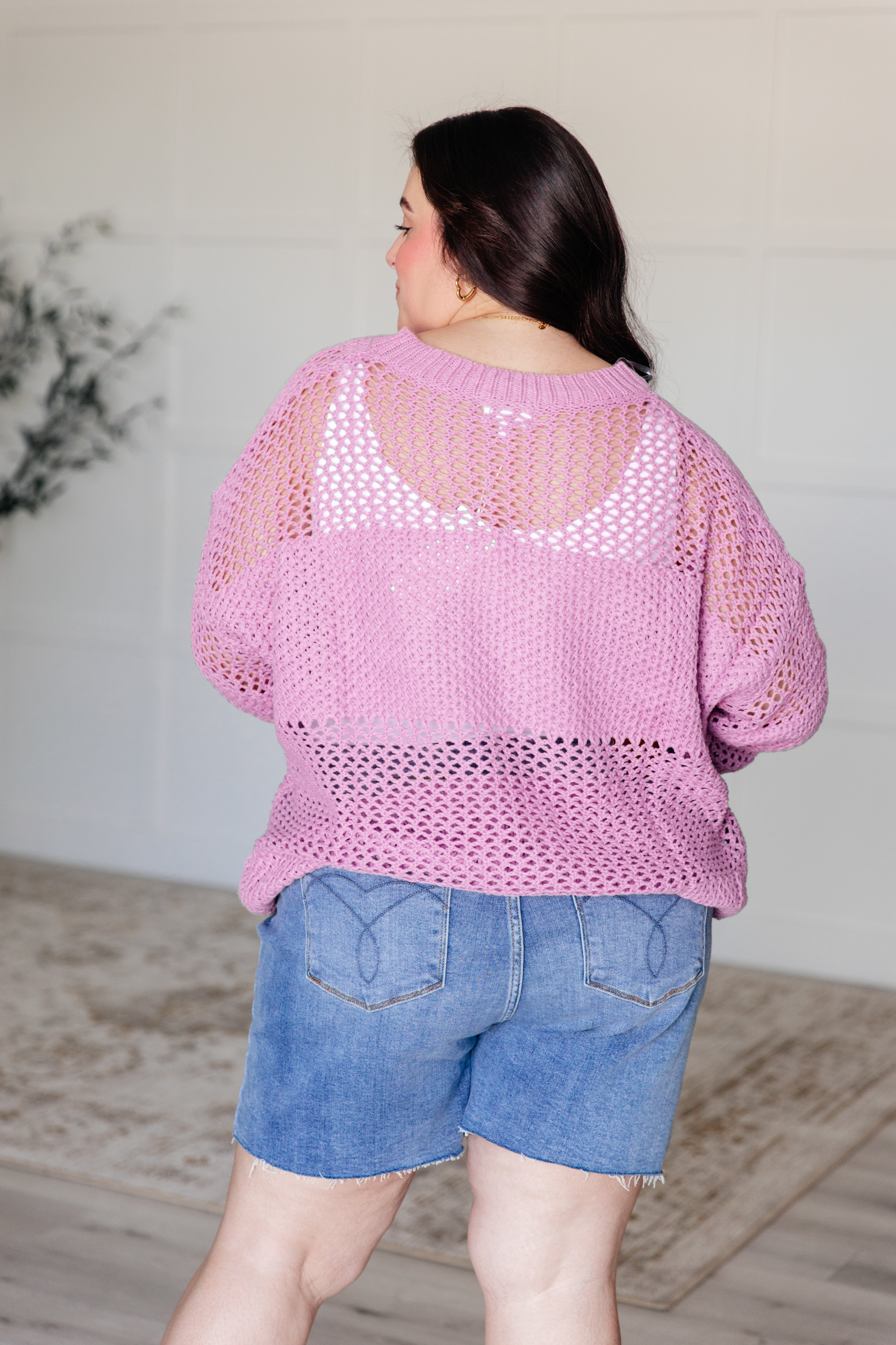 Woman seen from the back wearing a loose knit lavender sweater over a white top paired with denim shorts indoors