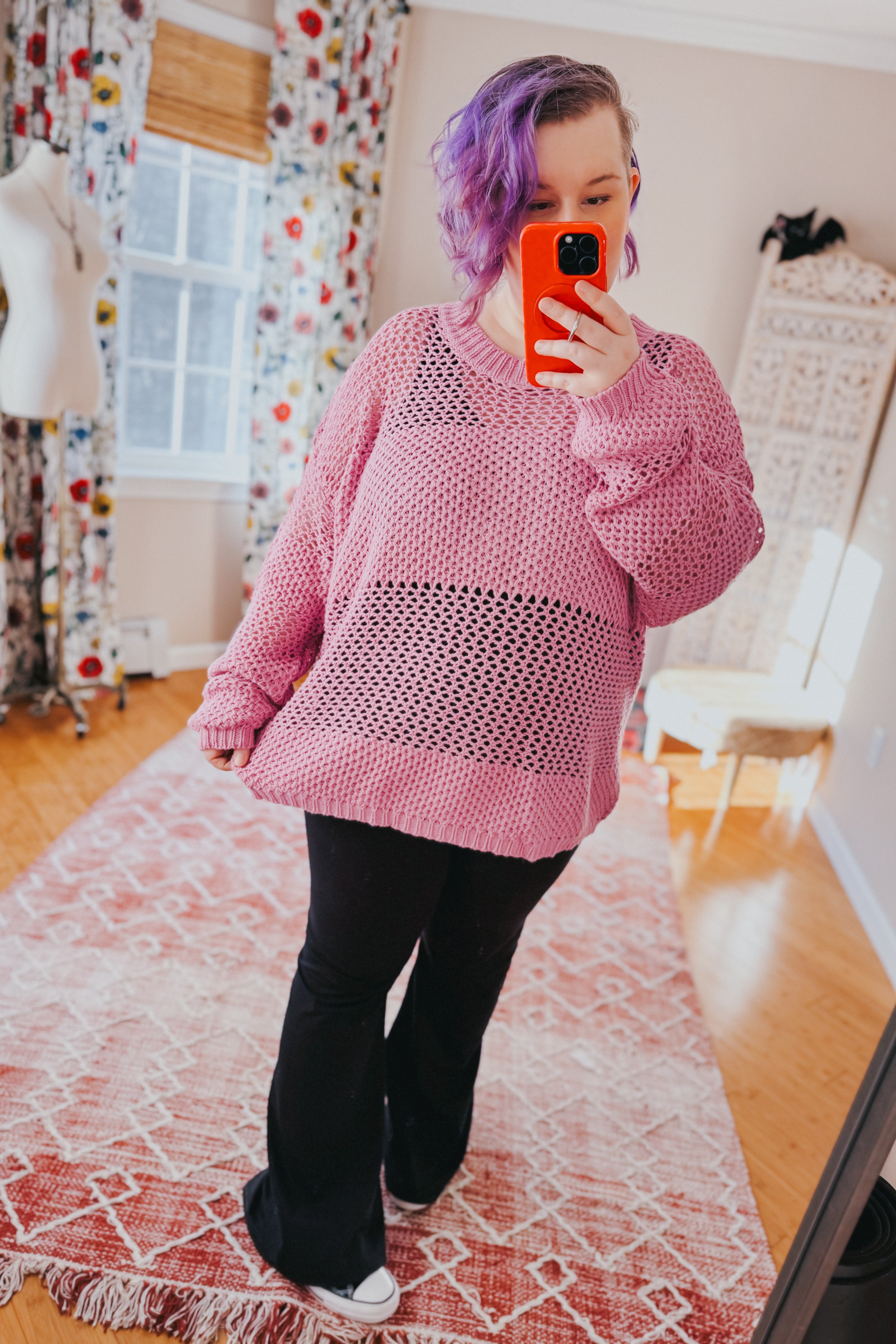 Woman with purple hair taking a mirror selfie wearing a loose pink open-knit sweater and black pants standing indoors