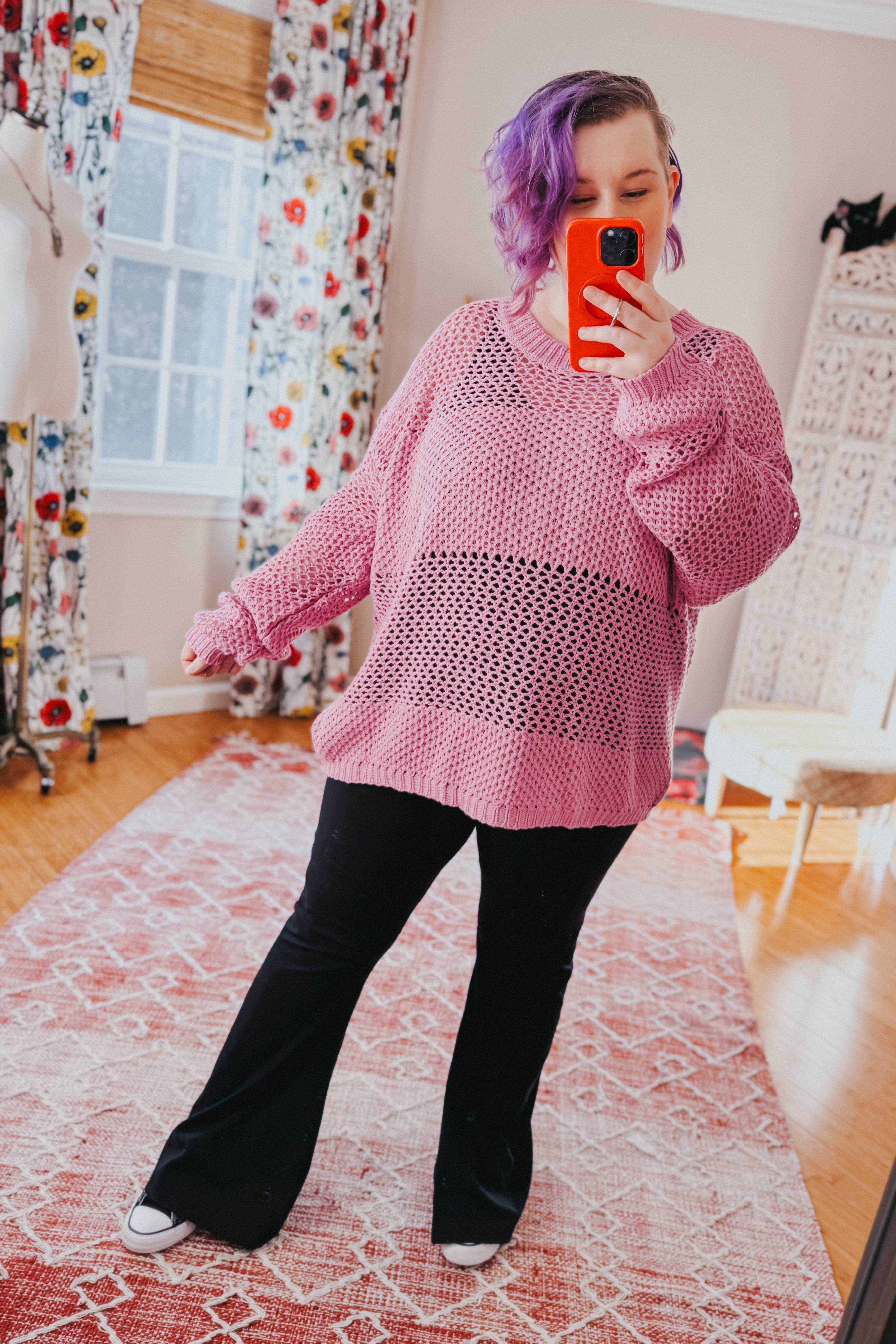 Woman with purple hair taking a mirror selfie wearing a loose pink open-knit sweater and black flared pants indoors