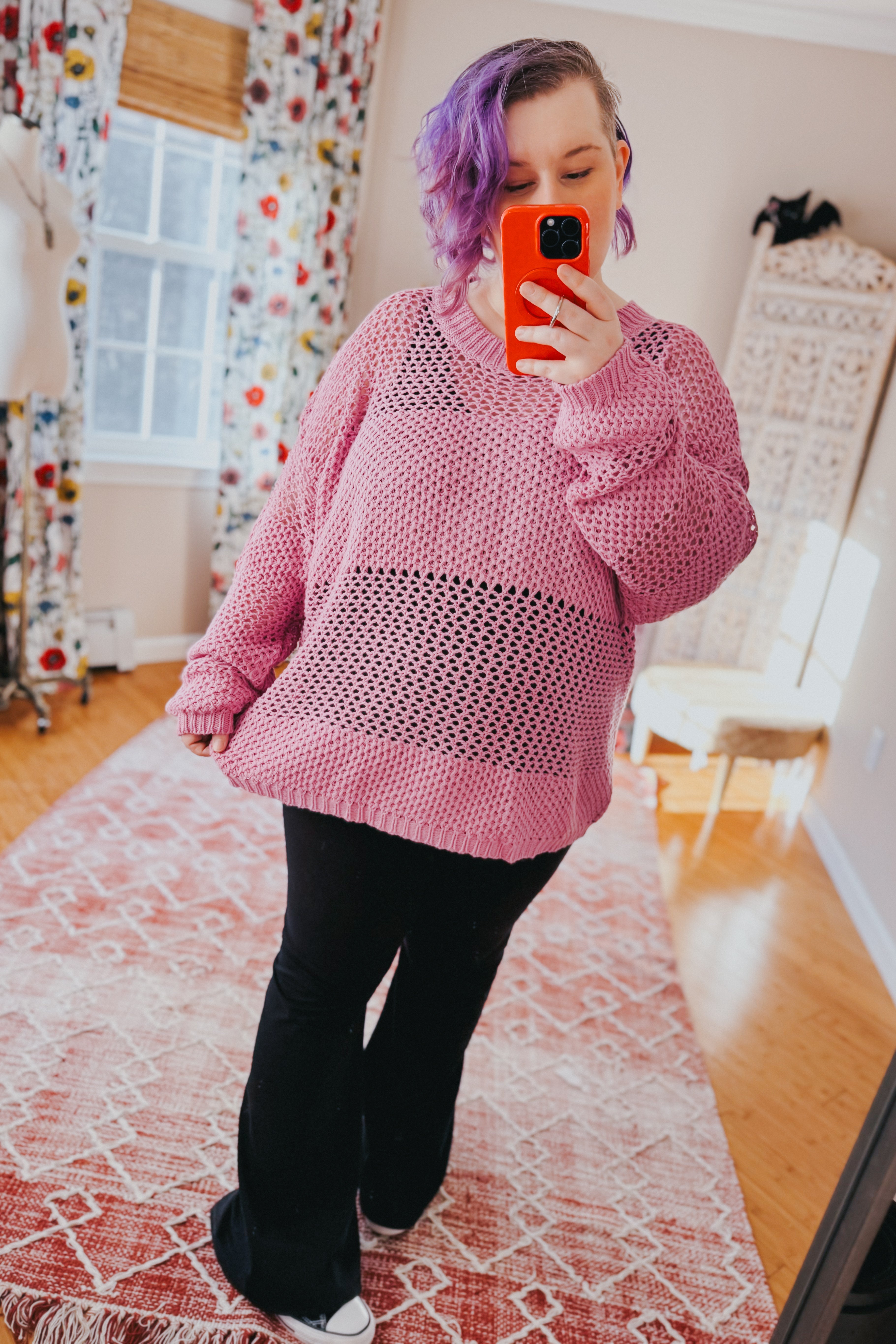 Woman with short purple hair taking a mirror selfie wearing a loose pink open-knit sweater and black pants indoors