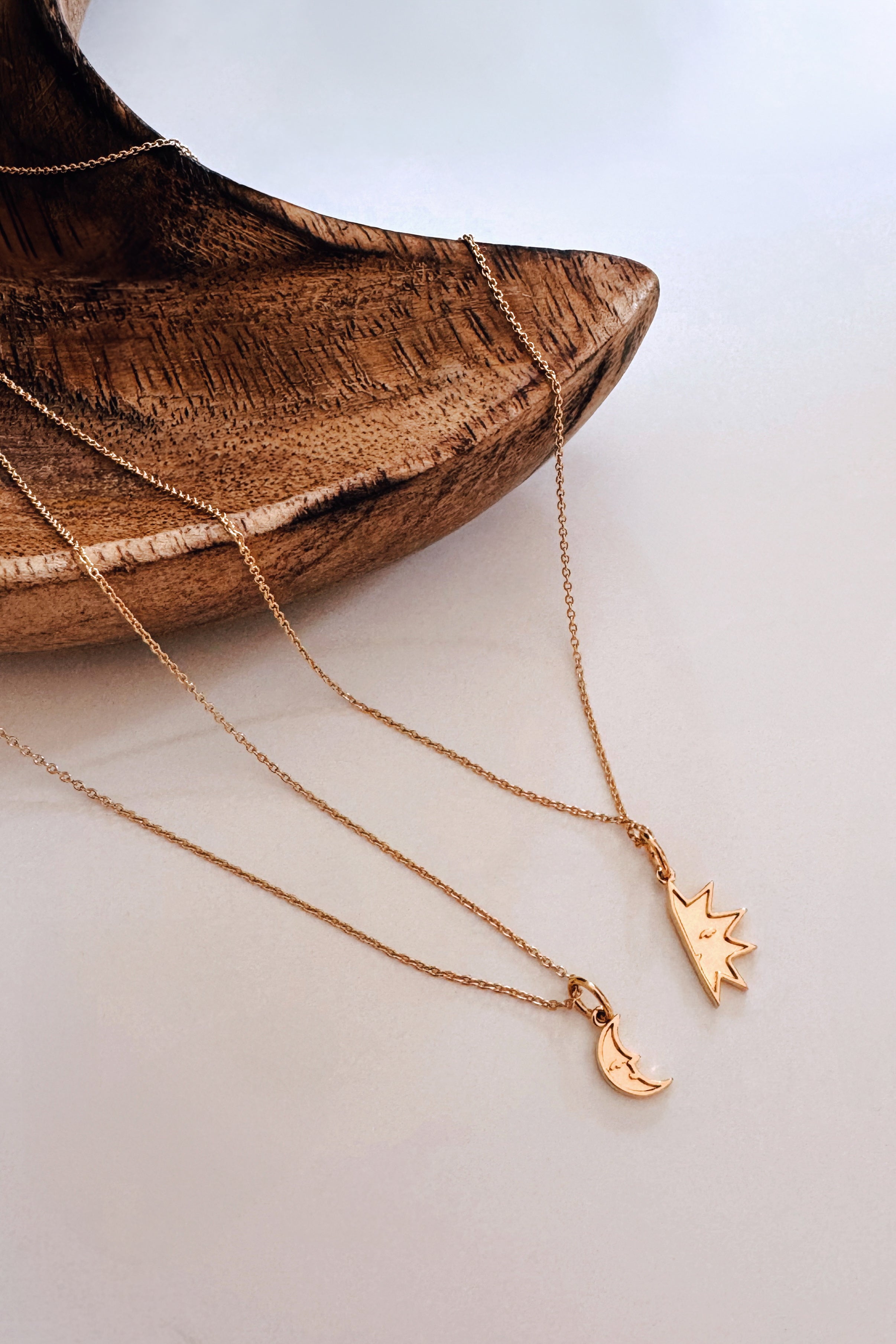 Two gold necklaces with small sun and crescent moon pendants displayed on a smooth white surface near a wooden bowl with natural grain.