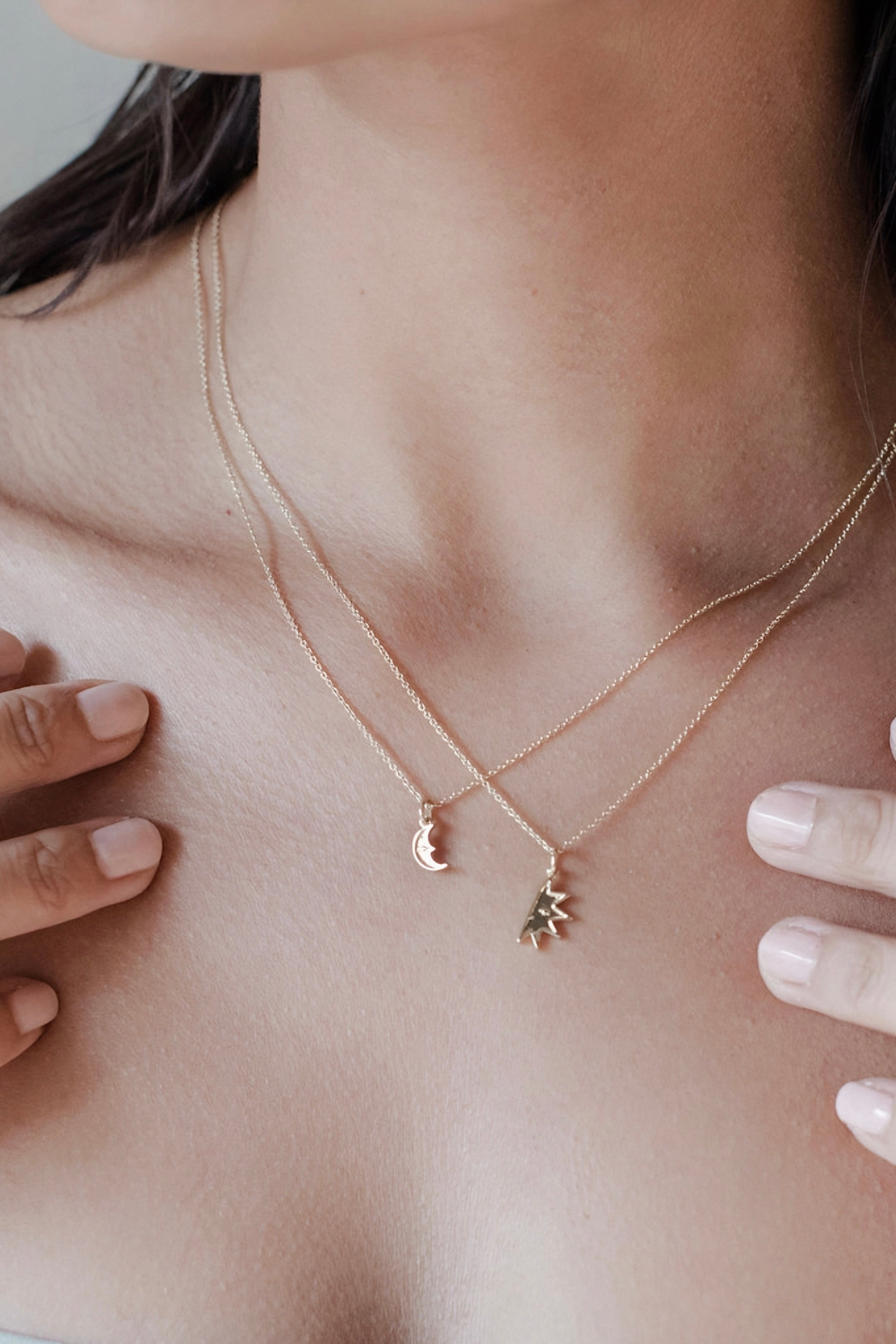 Close-up of a person wearing two delicate gold necklaces with sun and crescent moon pendants on fine chains against bare skin.