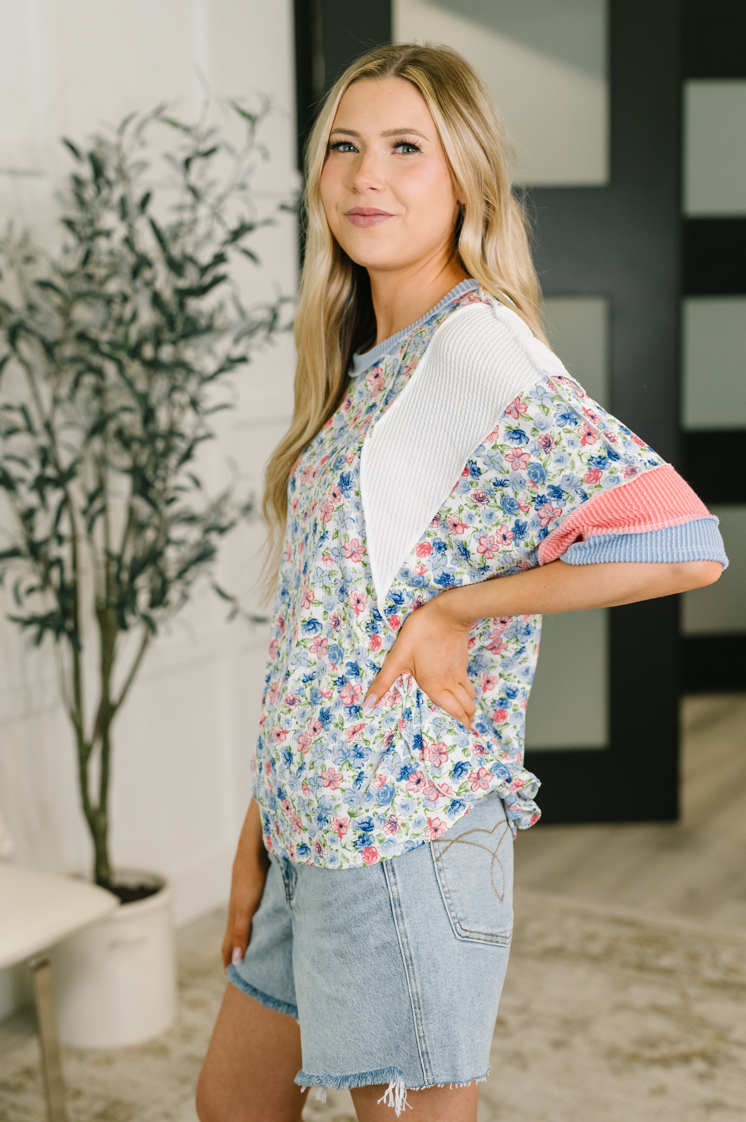 Side view of woman wearing a floral top with white and pink cable ribbed knit sleeves paired with denim shorts indoors
