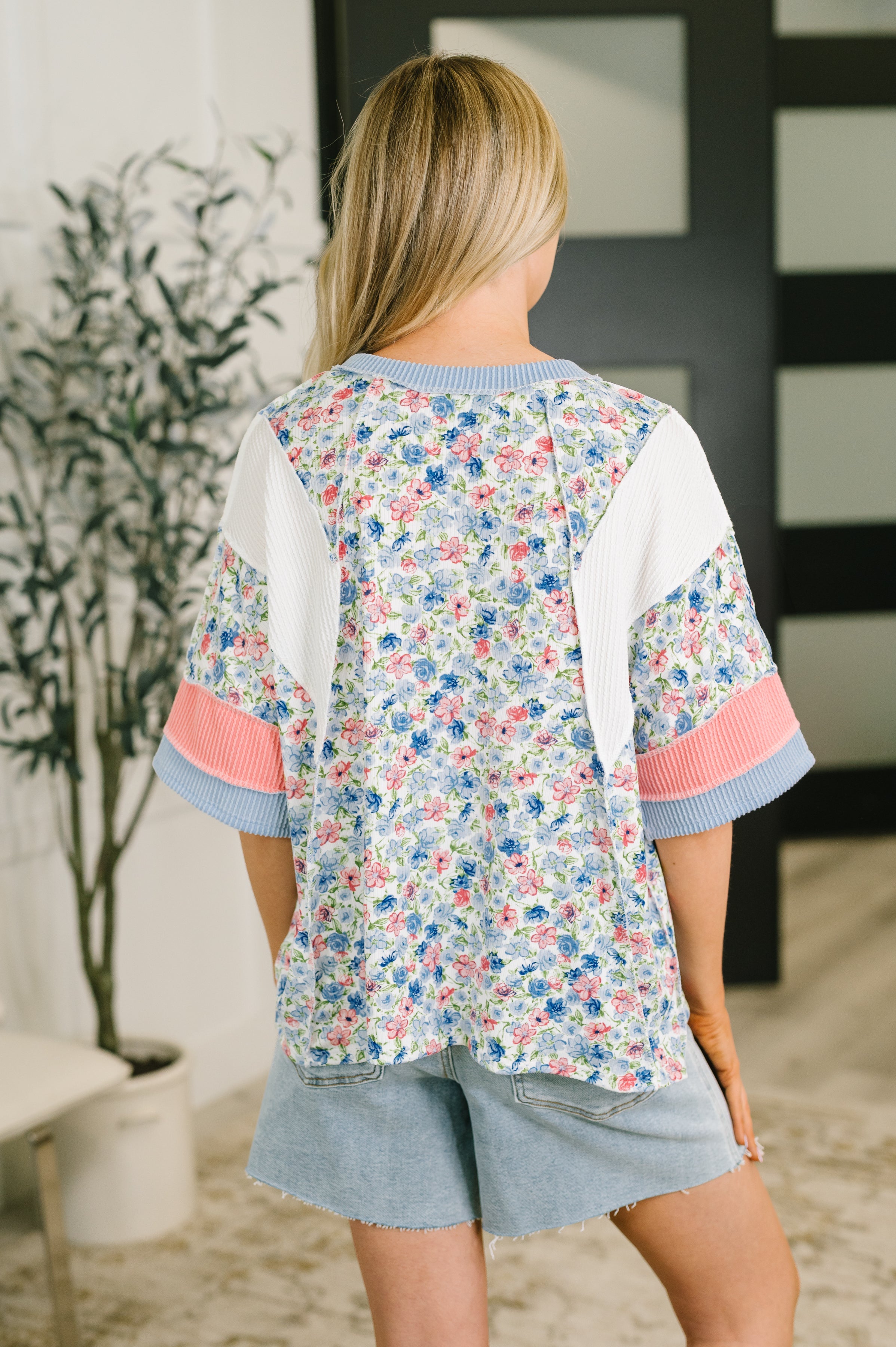 Back view of woman wearing a floral print top with ribbed white panels and pink and blue cuff accents indoors