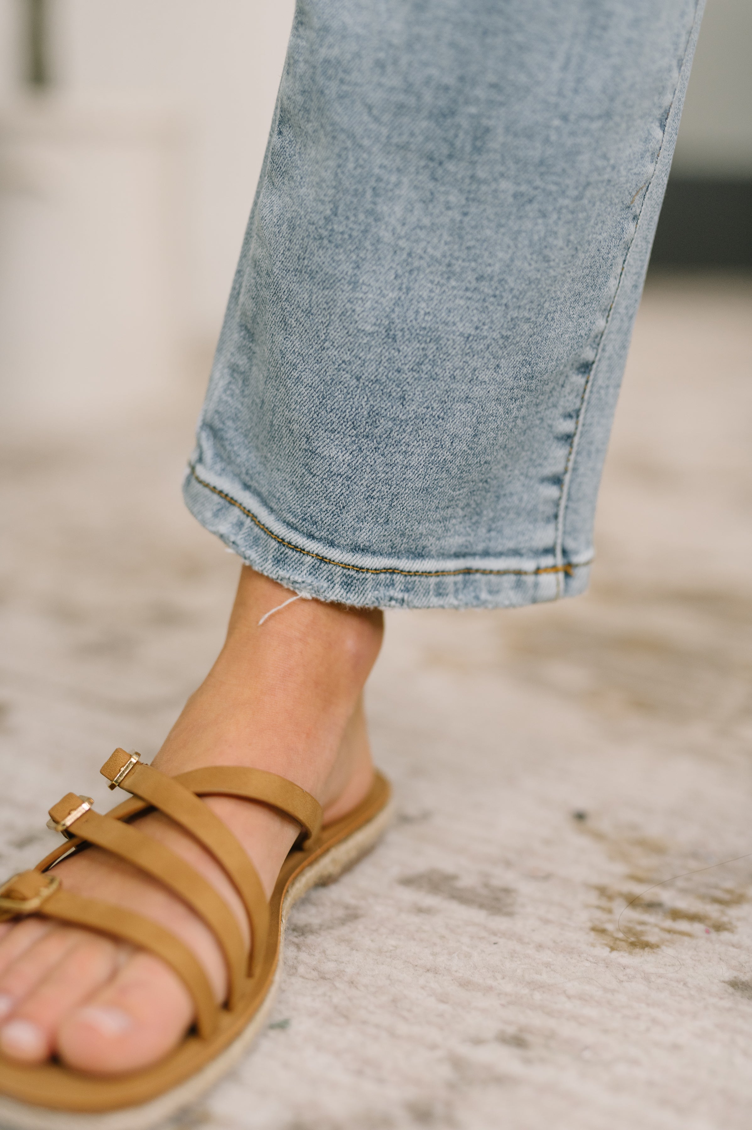 Close-up of raw hem detail on blue Natalie High Waist Ankle Straight Jeans paired with tan strappy sandals indoors