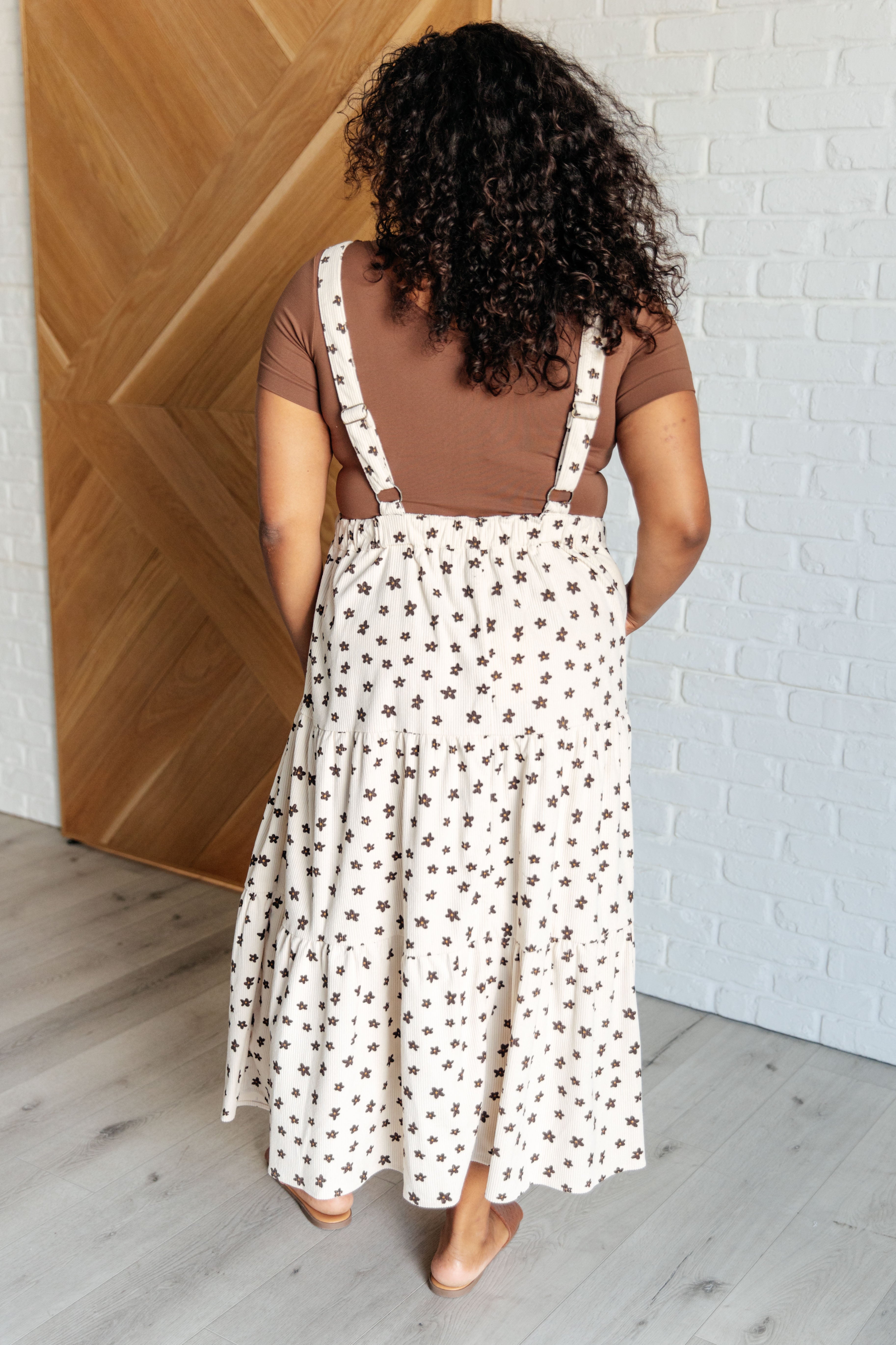 Woman with curly hair wearing a cream suspender skirt with small dark star prints and a brown short-sleeve top indoors