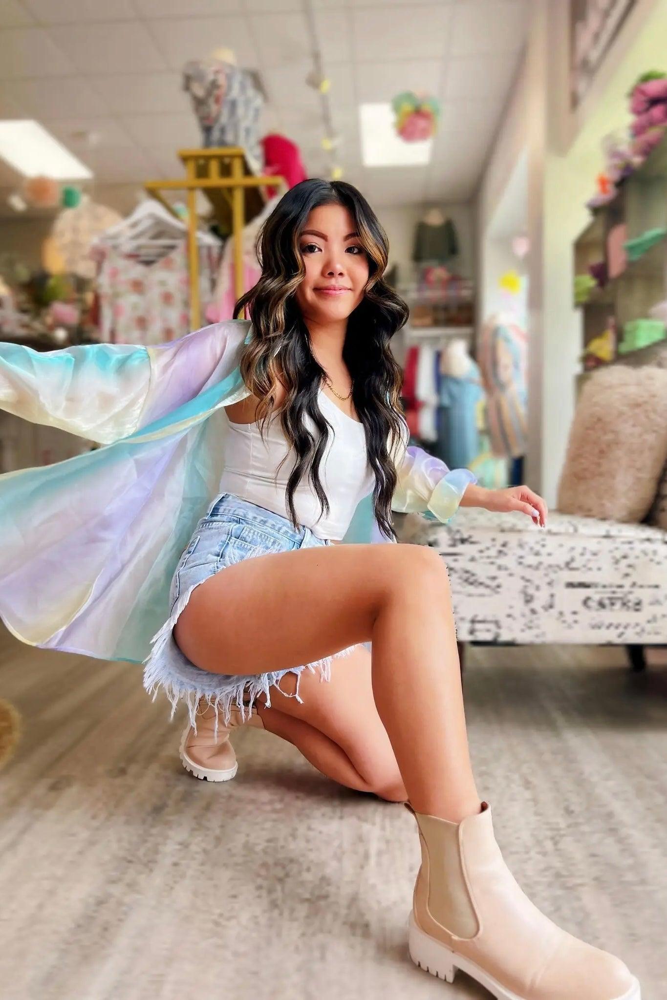 Woman posing indoors wearing a sheer blue and pastel iridescent button-up top over a white tank paired with light denim shorts and beige ankle boots.