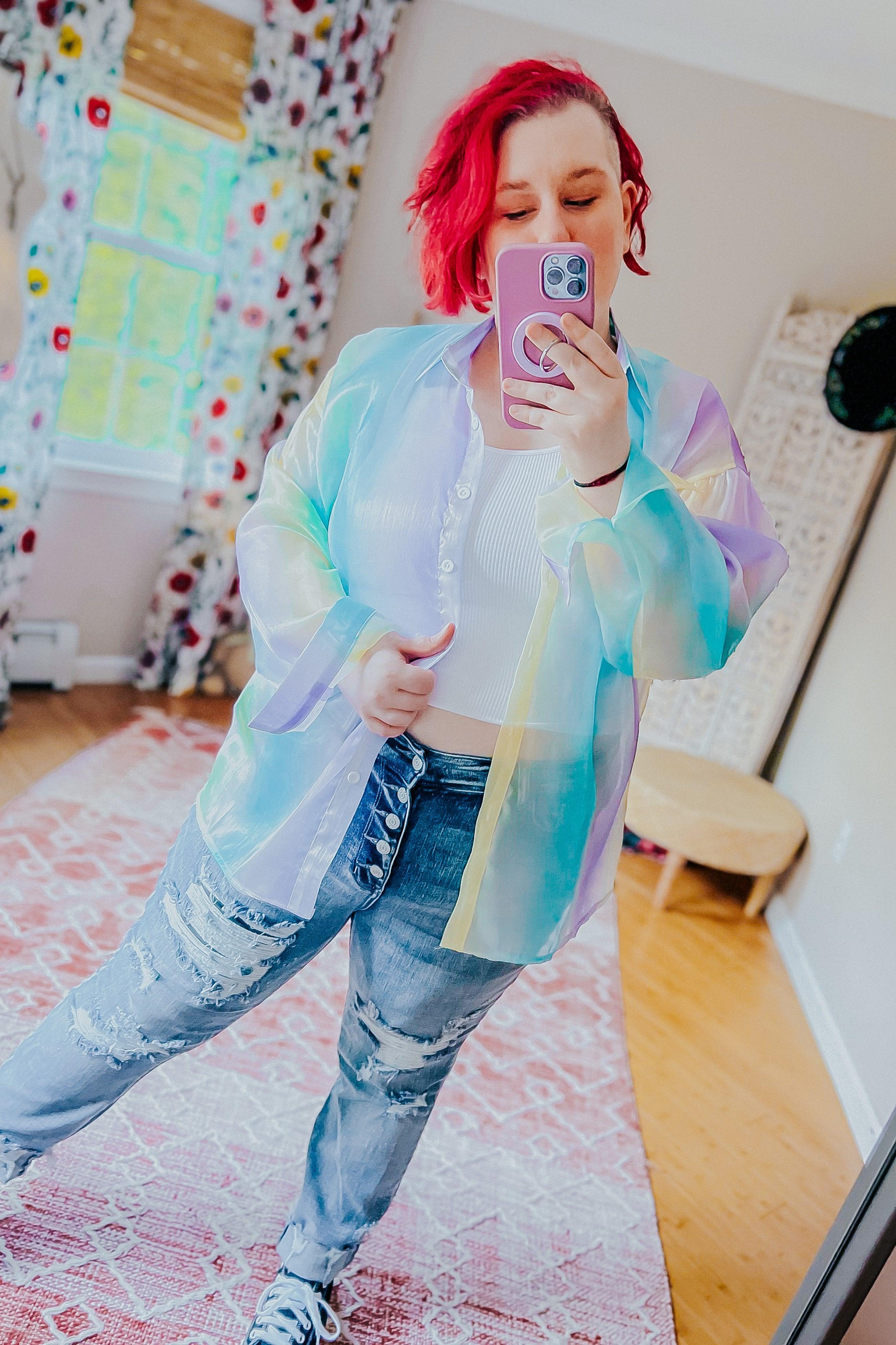 Woman wearing a sheer iridescent button-up top in pastel blue, green, and yellow tones over a white ribbed crop top with distressed high-waisted jeans in a bright, cozy room.