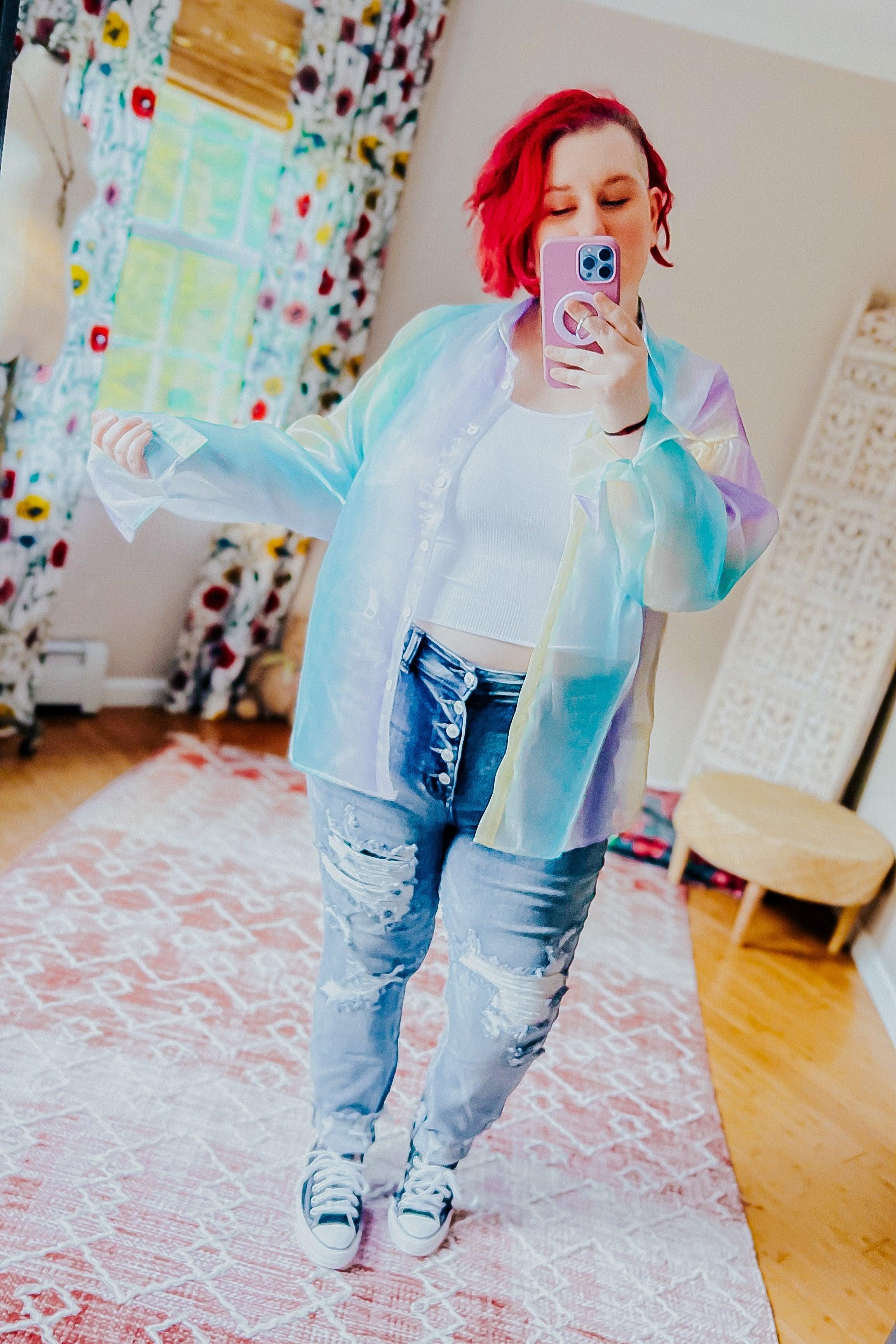 Woman taking a mirror selfie wearing a sheer blue iridescent button-up top over a white crop top with distressed high-rise jeans and sneakers in a bright room