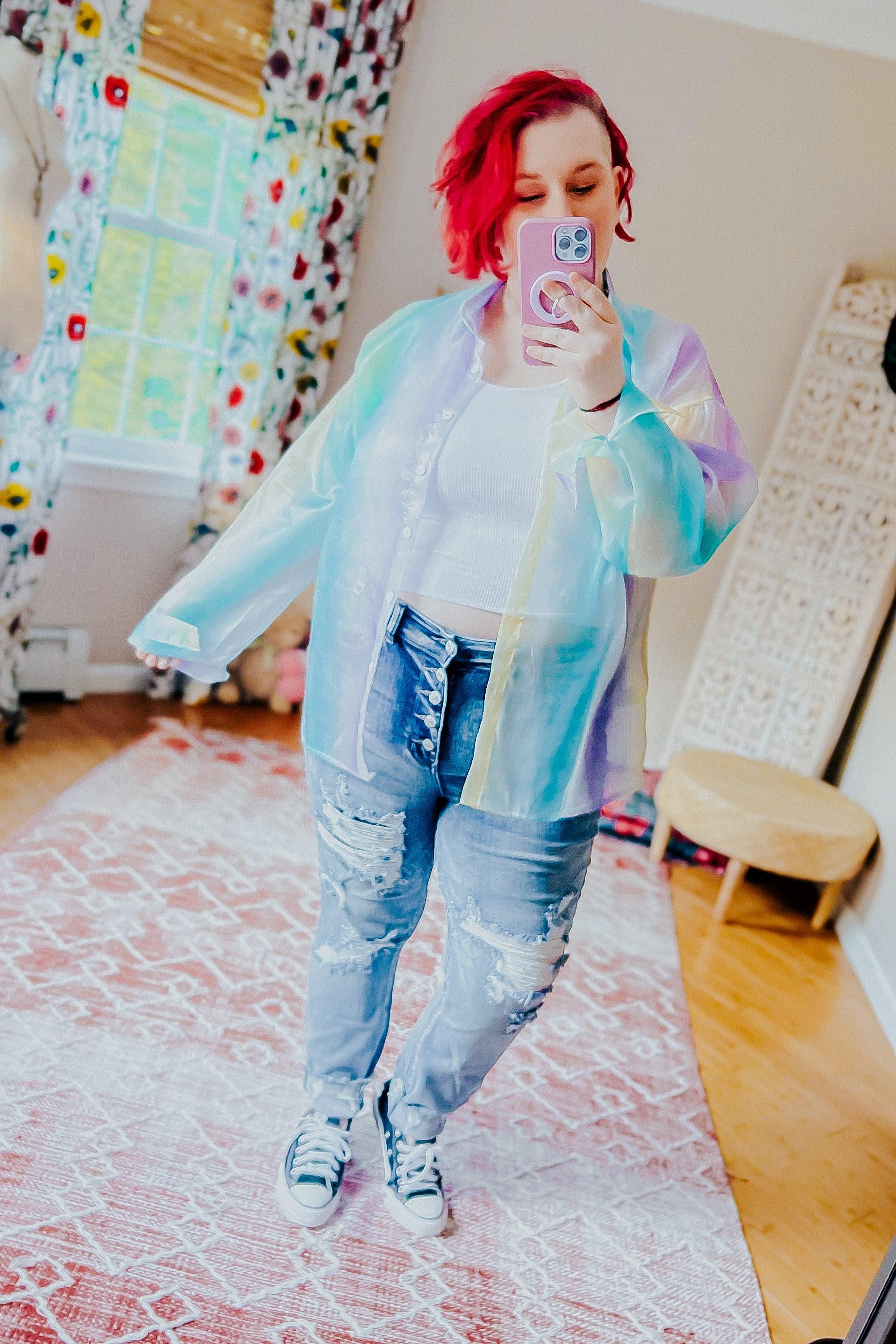 Woman taking a mirror selfie wearing a sheer, pastel blue and lavender iridescent button-up top over a white crop top with distressed high-rise jeans and sneakers in a bright room