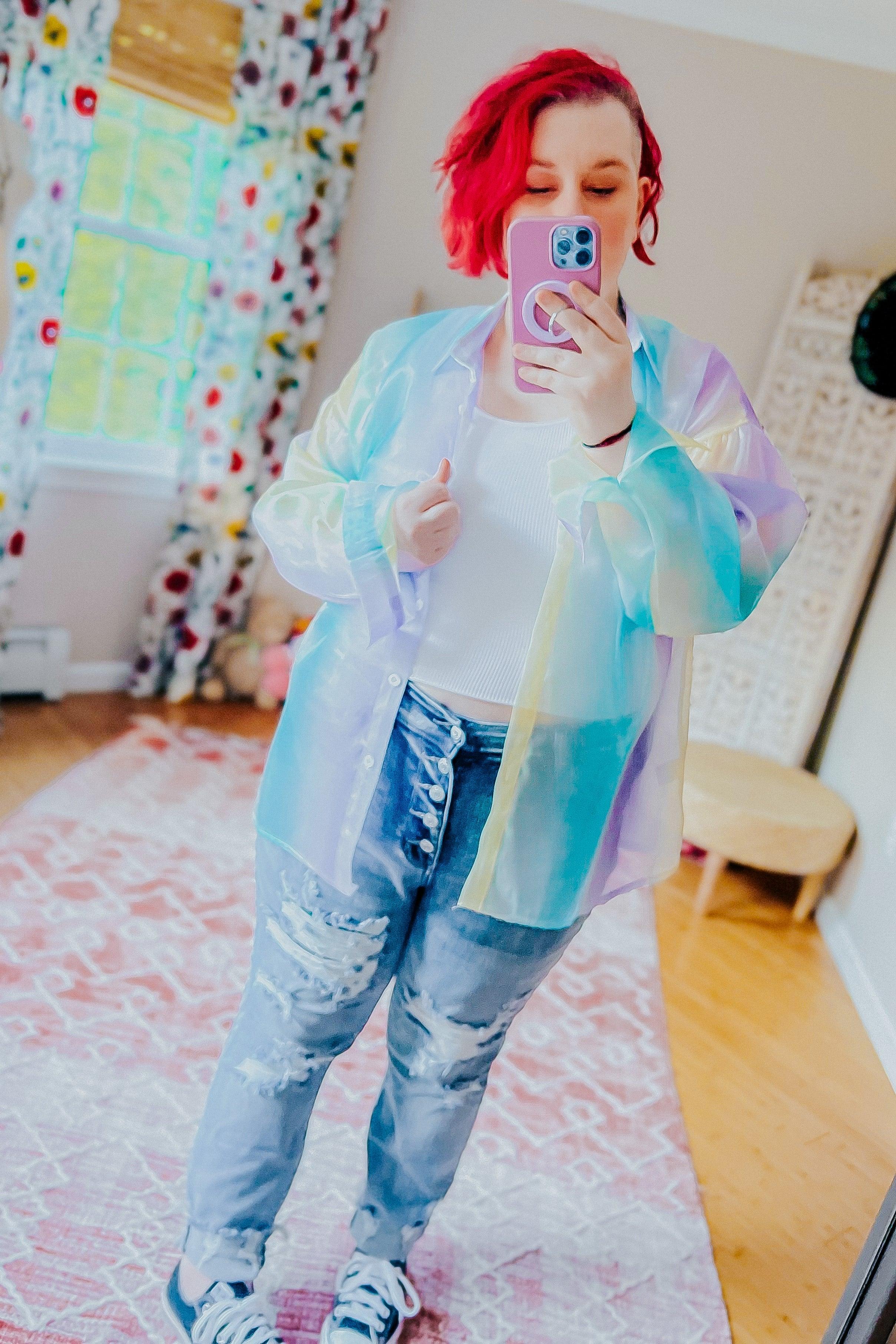 Woman taking a mirror selfie wearing a sheer, multicolored blue and pastel iridescent button-up top over a white ribbed tank with high-waisted distressed jeans and sneakers in a cozy room