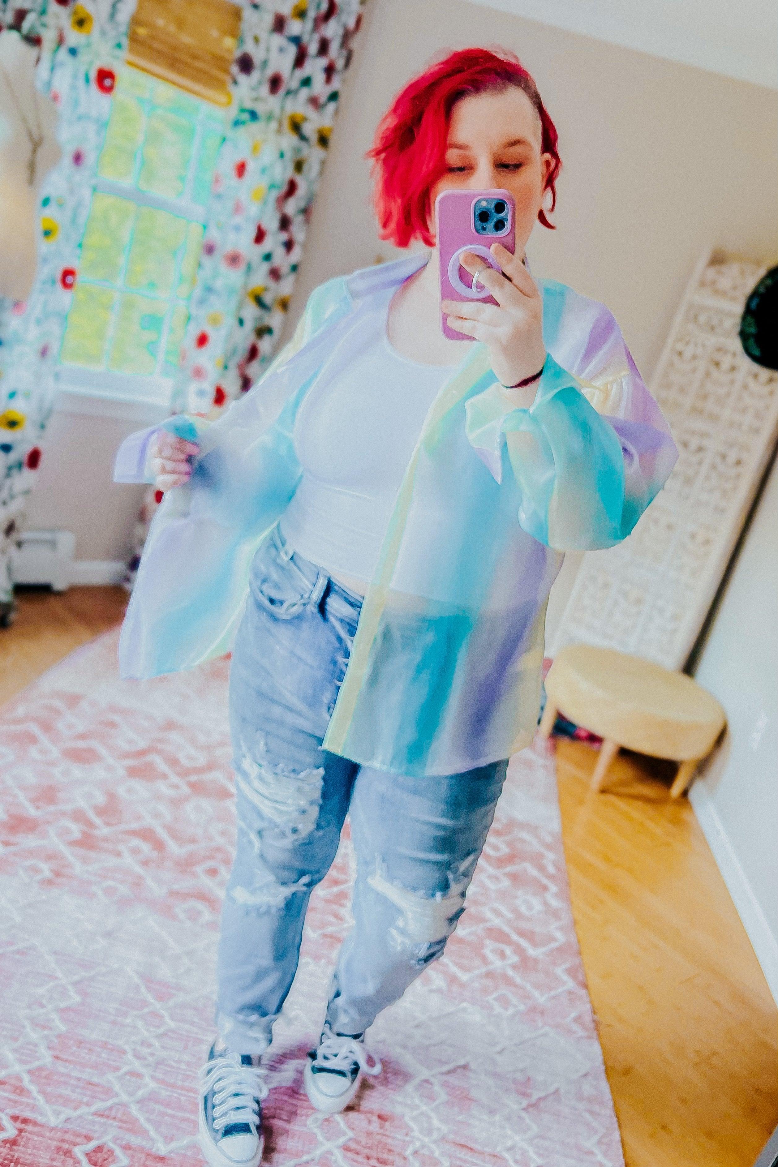 Woman taking a mirror selfie wearing a sheer, pastel blue and green iridescent button-up top over a white tank with light-wash distressed jeans and sneakers in a bright room