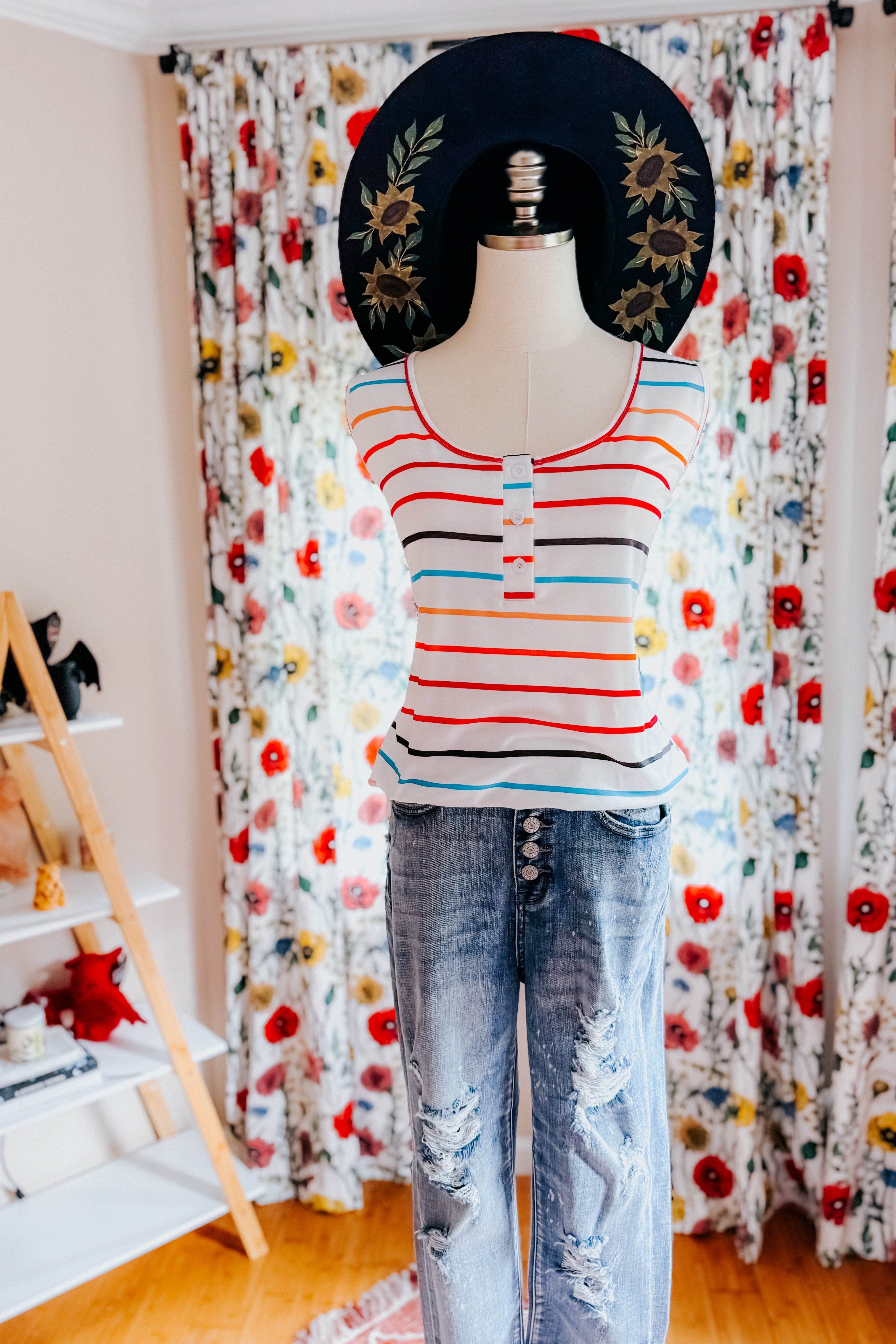 Mannequin wearing ivory striped sleeveless henley tank with blue distressed jeans and black wide-brim hat with floral design