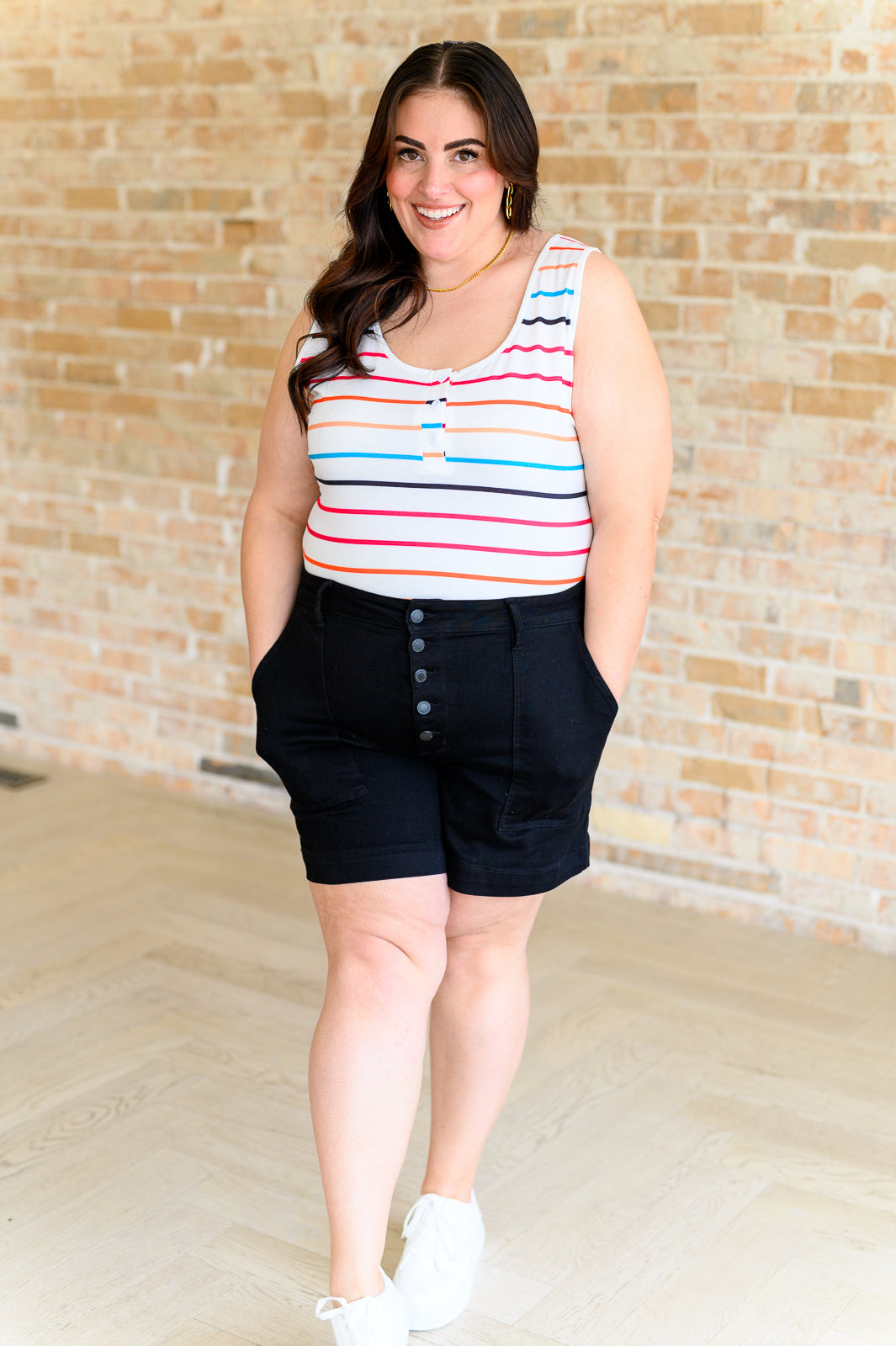 Plus size woman wearing colorful striped sleeveless tank top with button details and black high-waisted shorts indoors