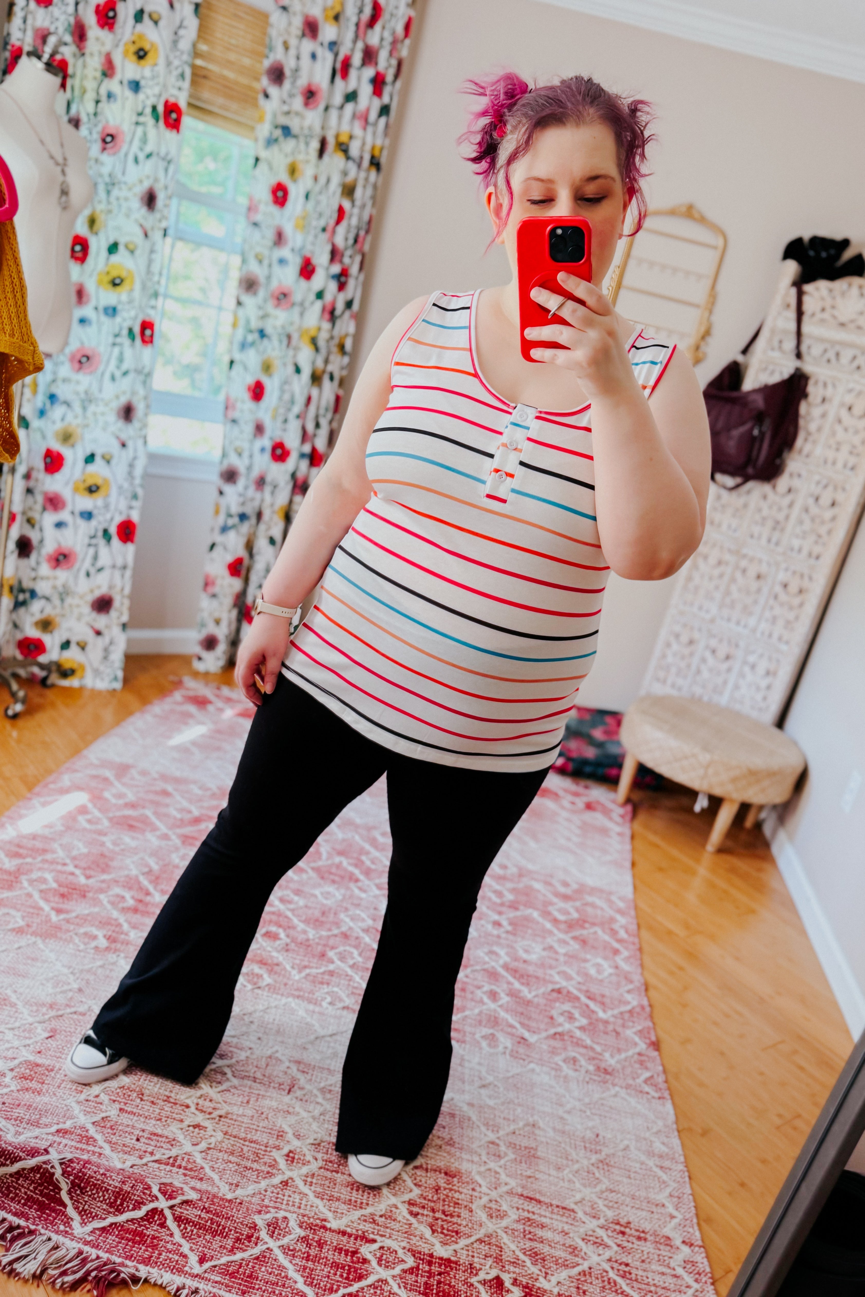 Woman taking mirror selfie wearing ivory sleeveless henley tank with colorful stripes and black pants in bright room