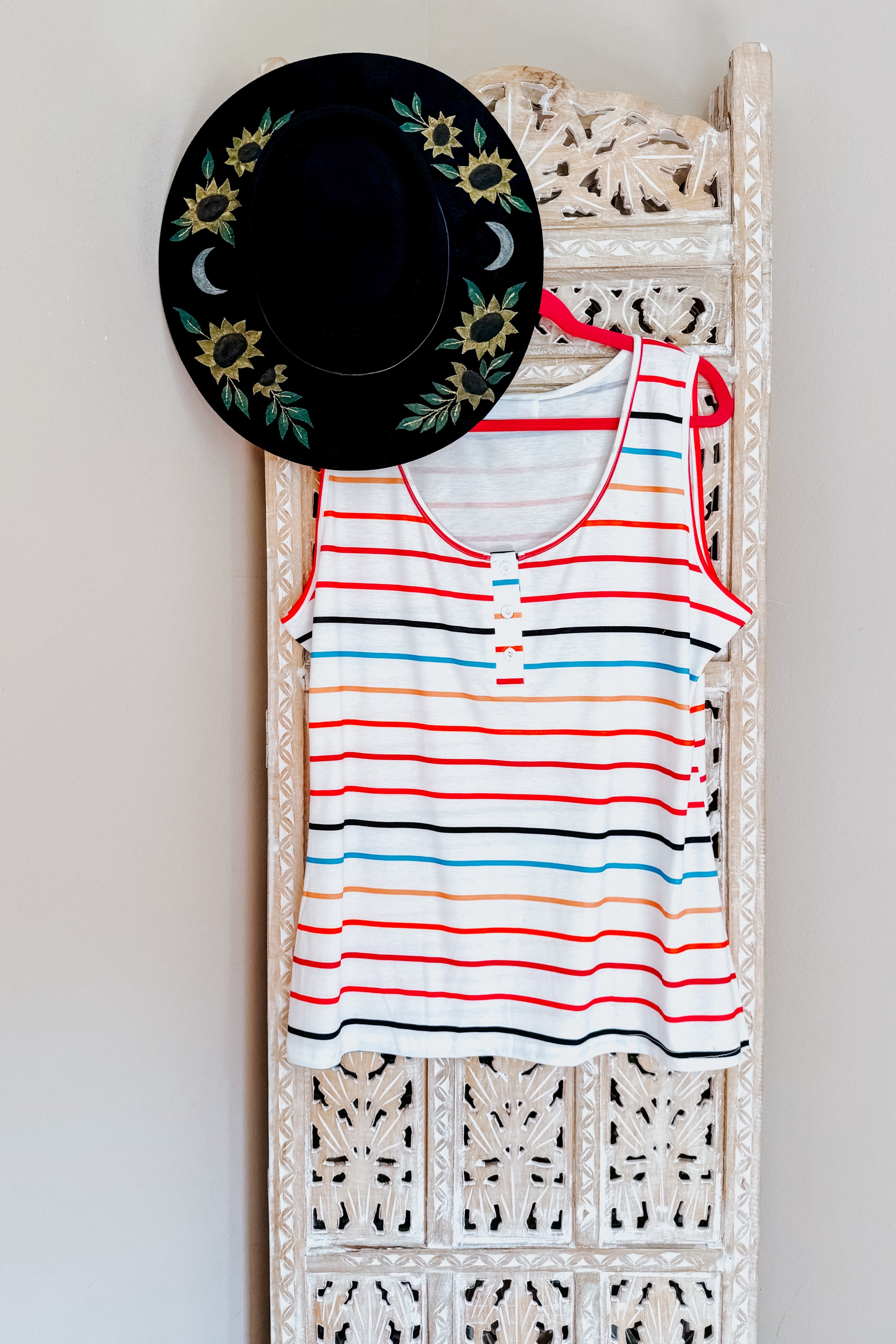 Ivory tank top with colorful horizontal stripes hanging on carved wooden panel paired with black floral hat