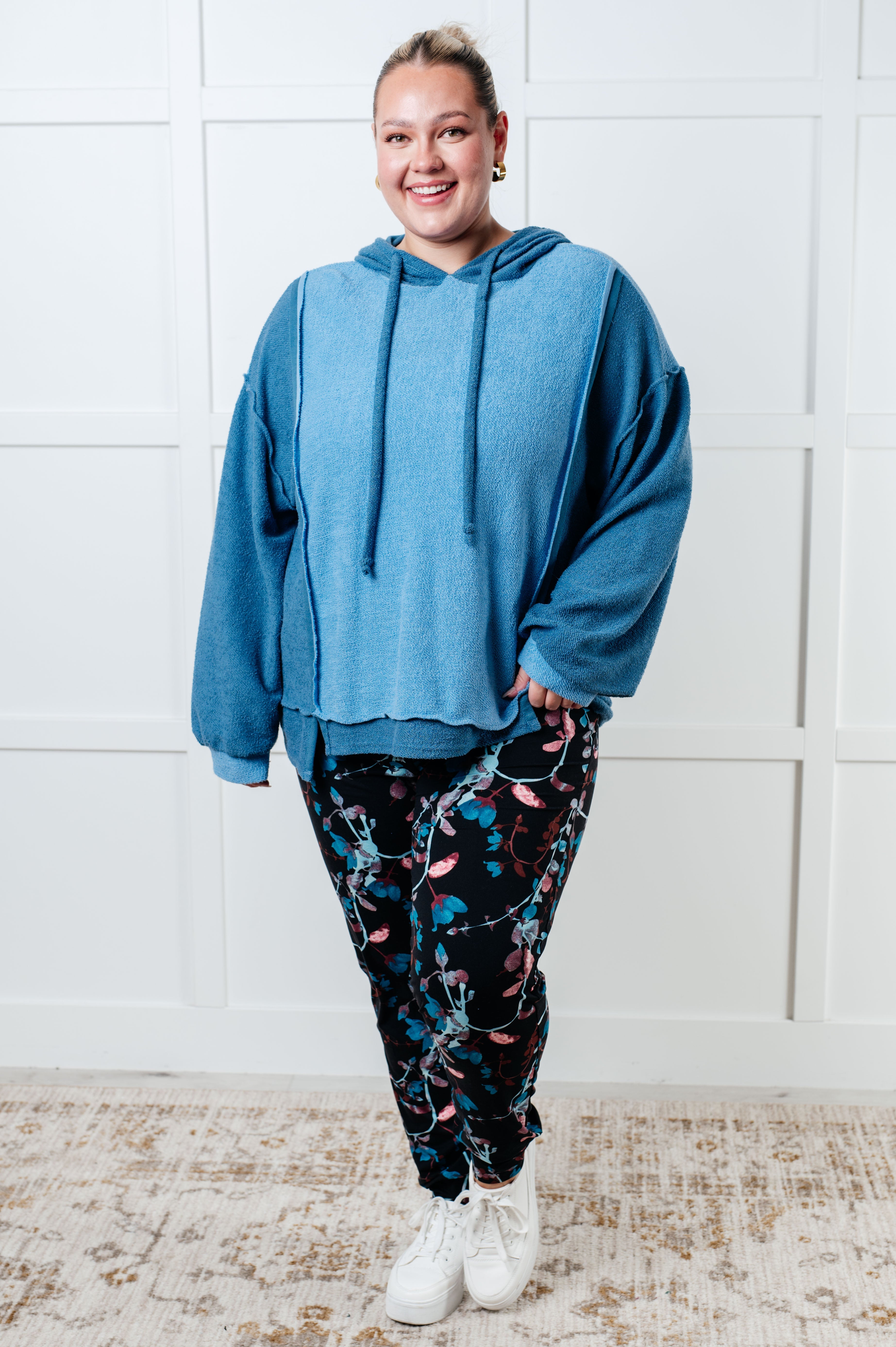 Smiling woman wearing a relaxed fit blue color block hoodie with long sleeves paired with floral patterned leggings and white sneakers indoors.