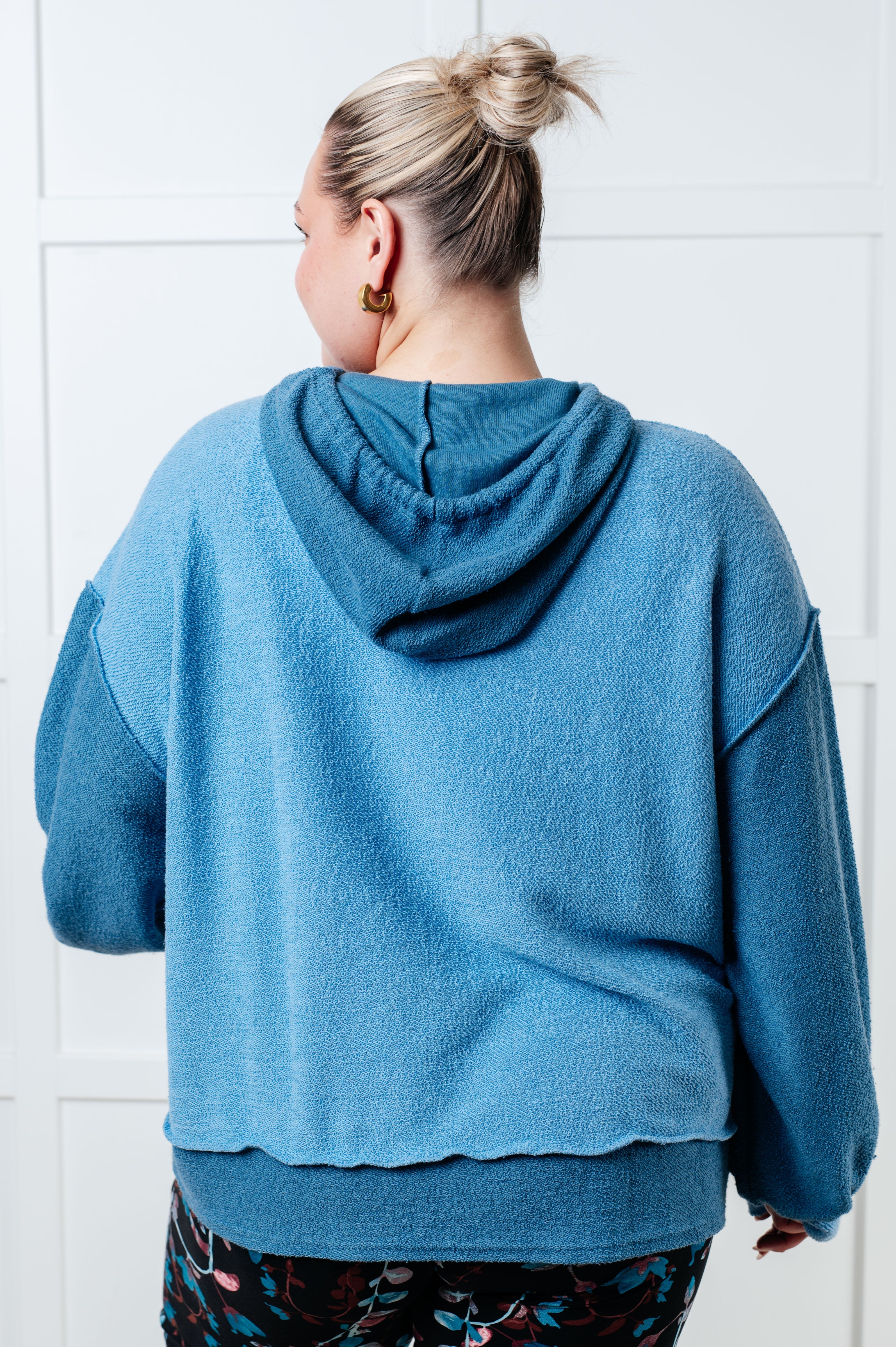 Woman with blonde hair in a top bun wearing a relaxed fit blue color block hoodie with a textured fabric, shown from the back indoors.