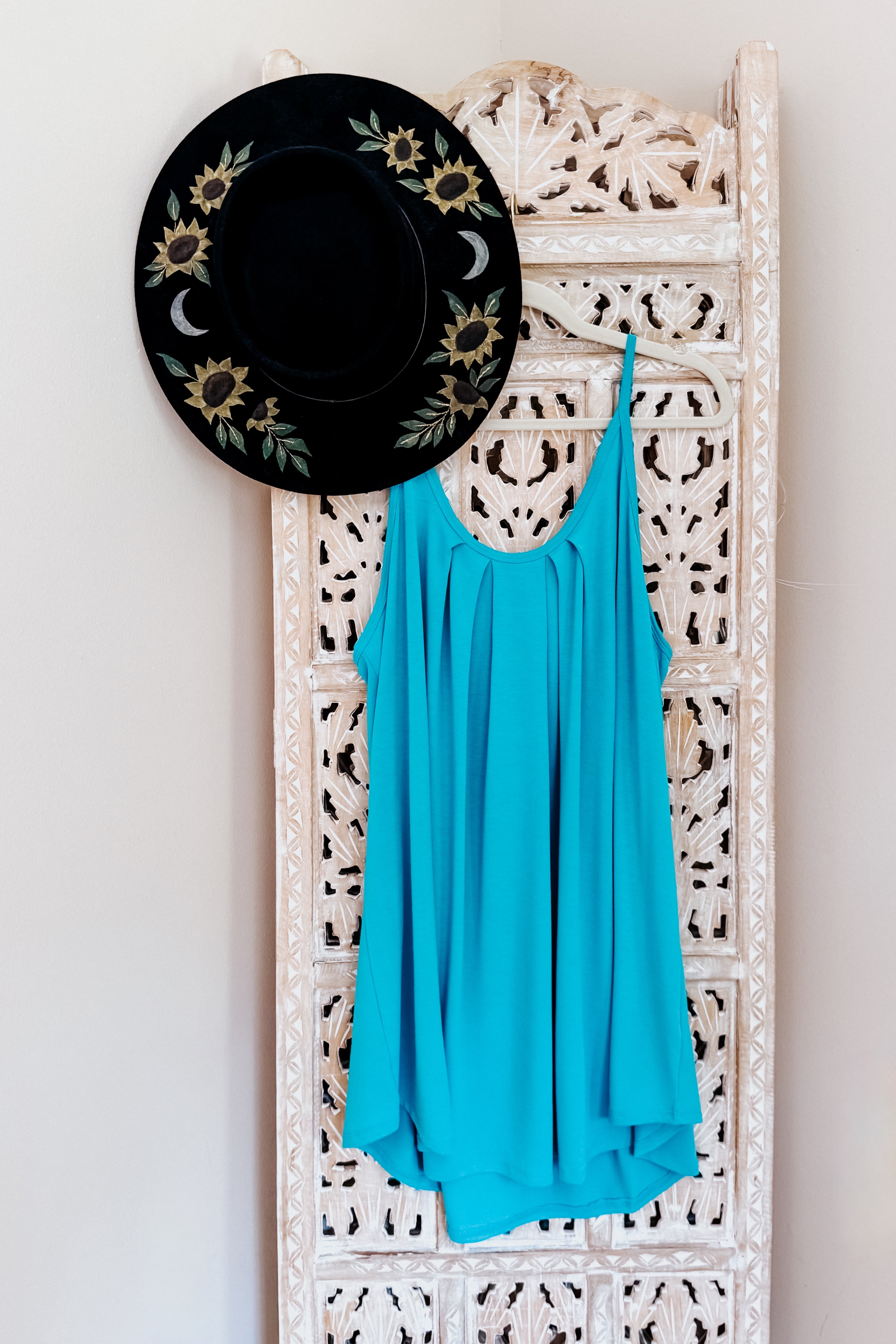 Bright neon blue pleated tank with thin straps hanging on a carved wooden panel next to a black hat decorated with sunflowers and crescent moons.