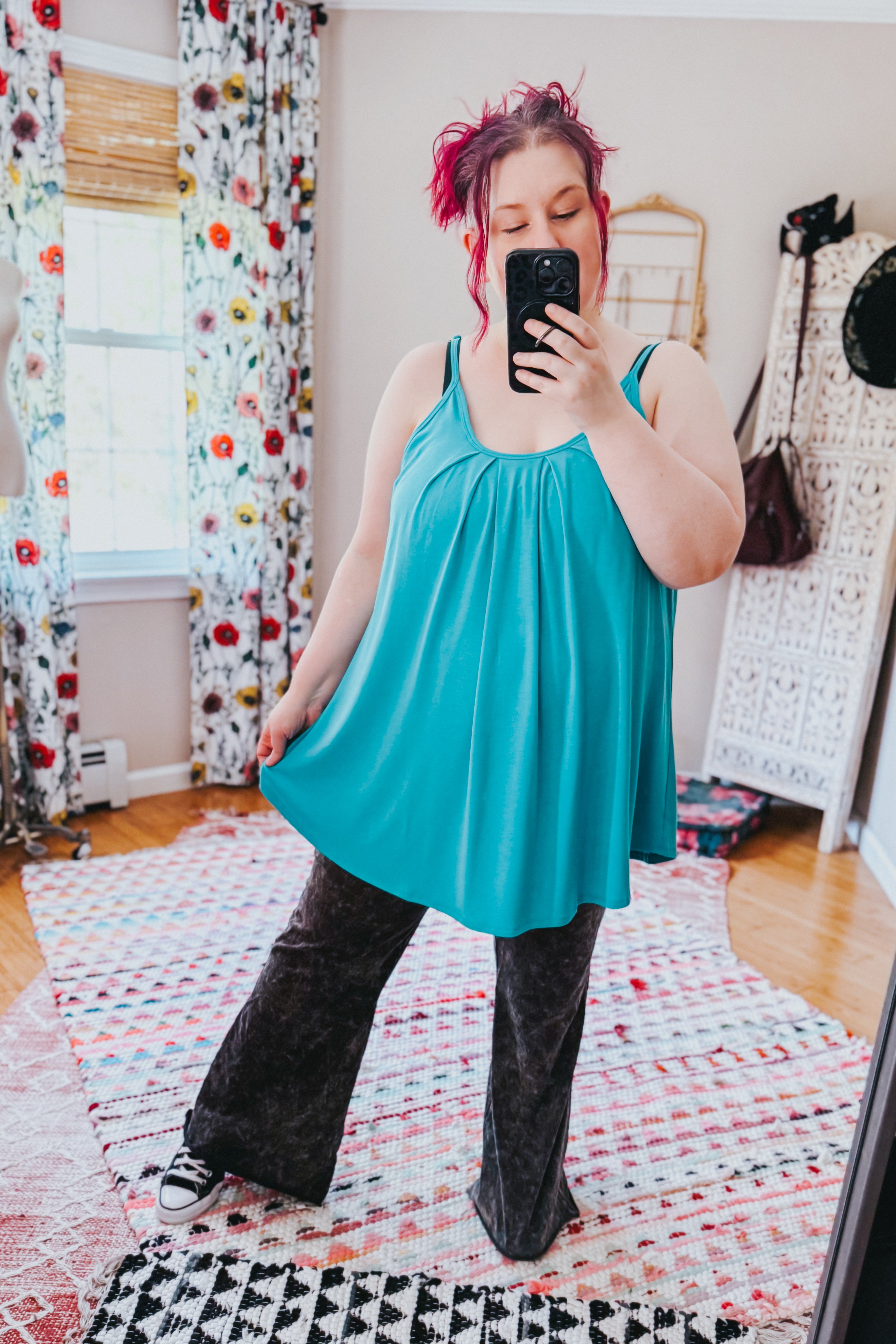 Woman taking a mirror selfie wearing a loose neon blue pleated tank top with thin straps paired with wide-leg black pants in a cozy room with patterned curtains and rugs.