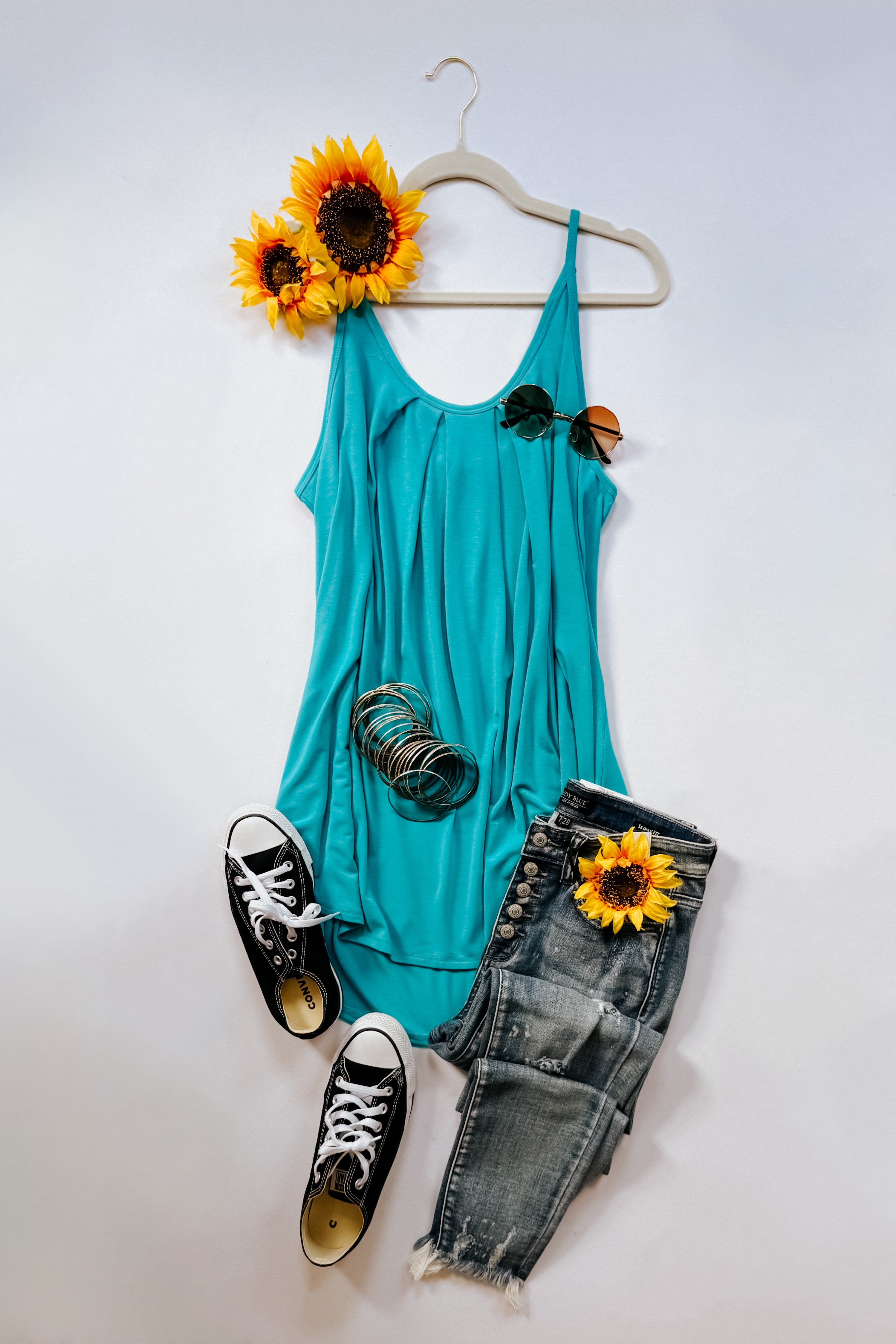 Bright neon blue pleated tank top with thin straps displayed on a white hanger, paired with distressed denim jeans, black sneakers, and sunflower accents.