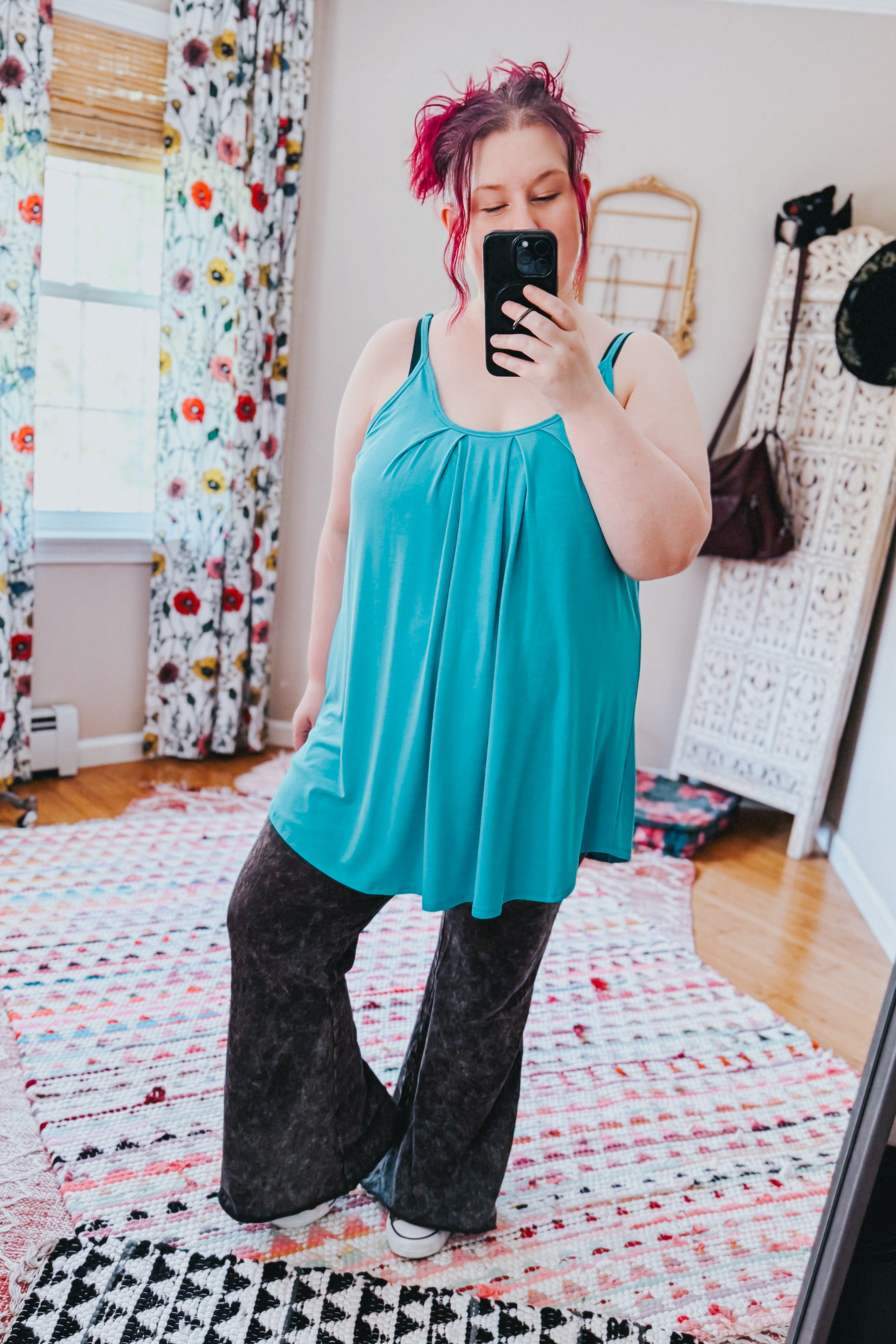 Woman taking a mirror selfie wearing a loose neon blue pleated tank top with thin straps paired with dark flared pants in a bright room with patterned curtains and rug.