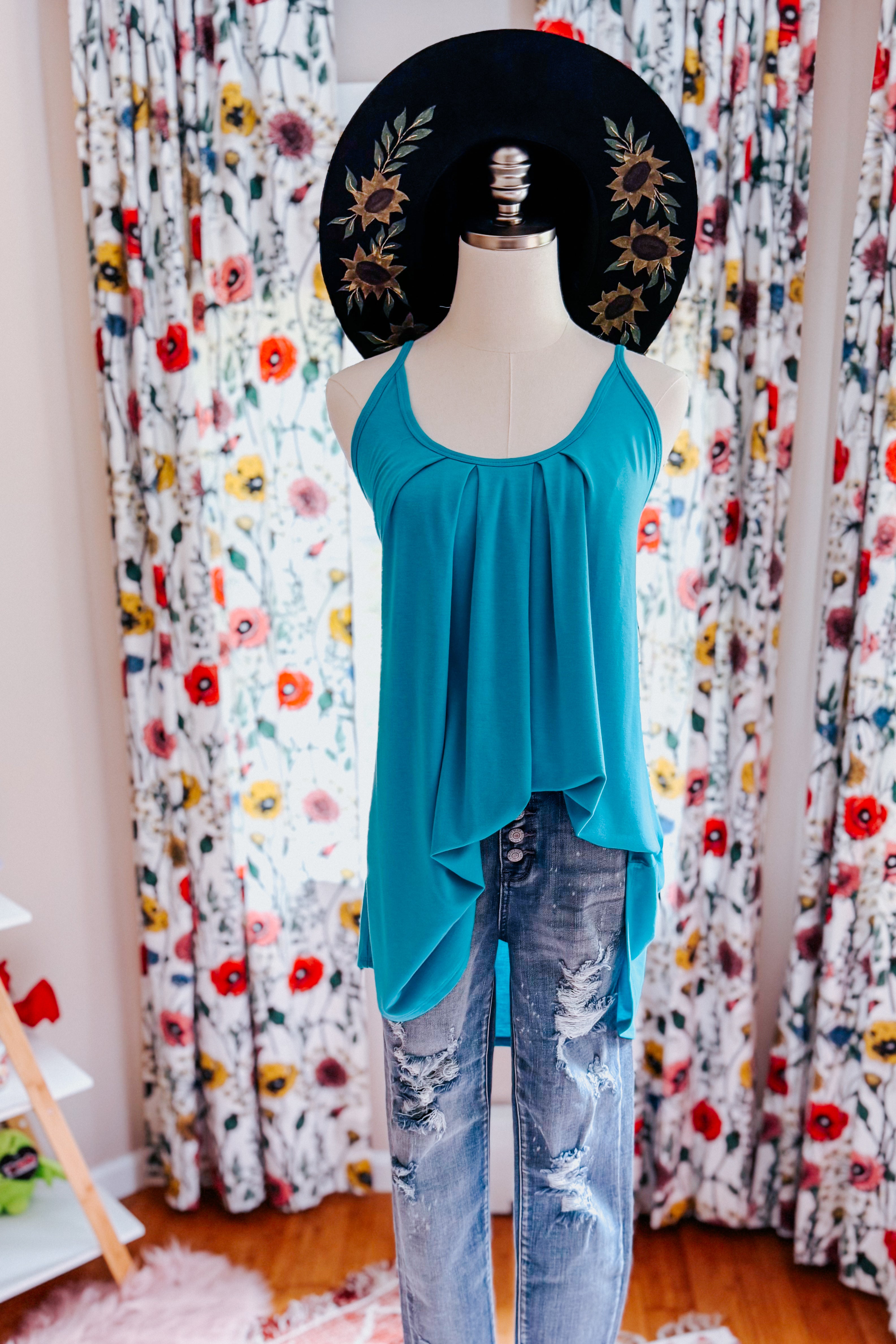 Mannequin dressed in a loose neon blue pleated tank top paired with distressed blue jeans, with a black wide-brim hat featuring sunflower embroidery, set against floral curtains.