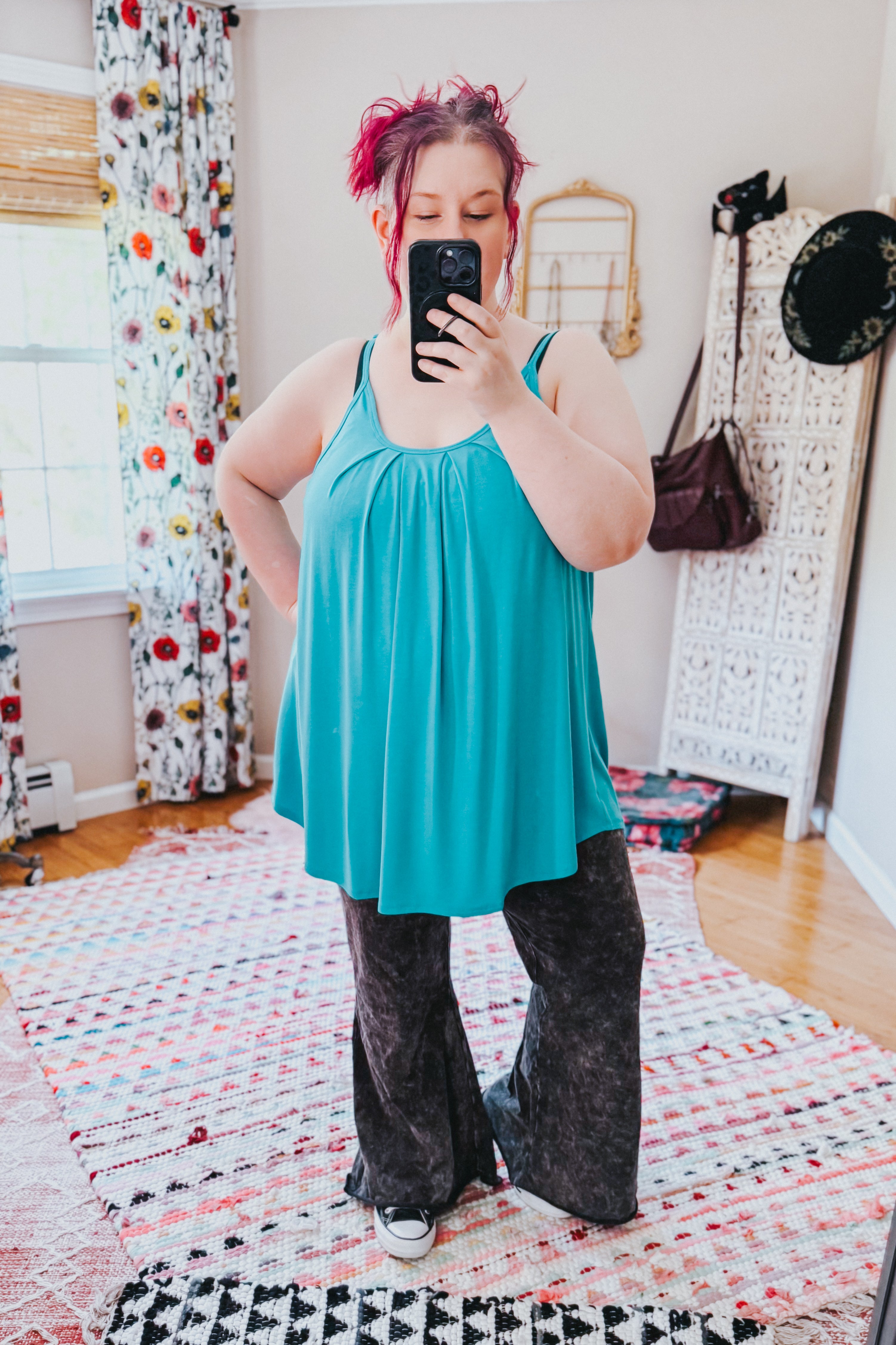 Woman taking a mirror selfie wearing a loose neon blue pleated tank top with thin straps paired with dark wide-leg pants in a bright room with floral curtains and patterned rugs.