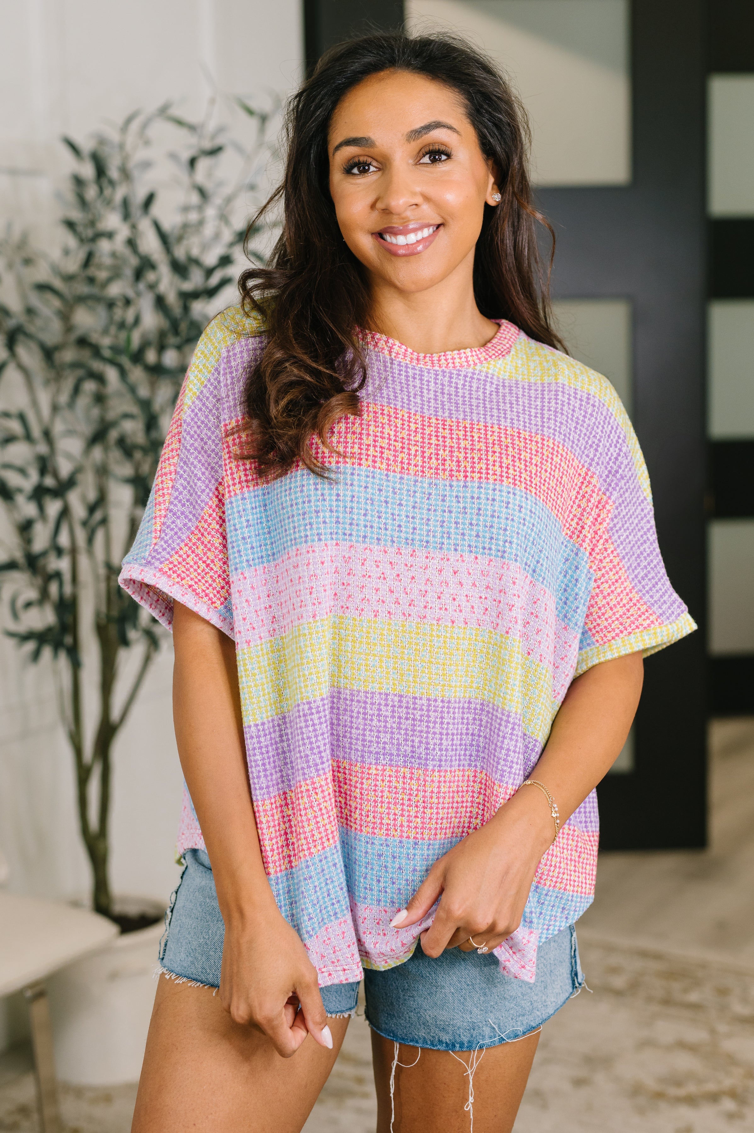 Smiling woman wearing pastel rainbow striped textured knit top with short sleeves and denim shorts indoors