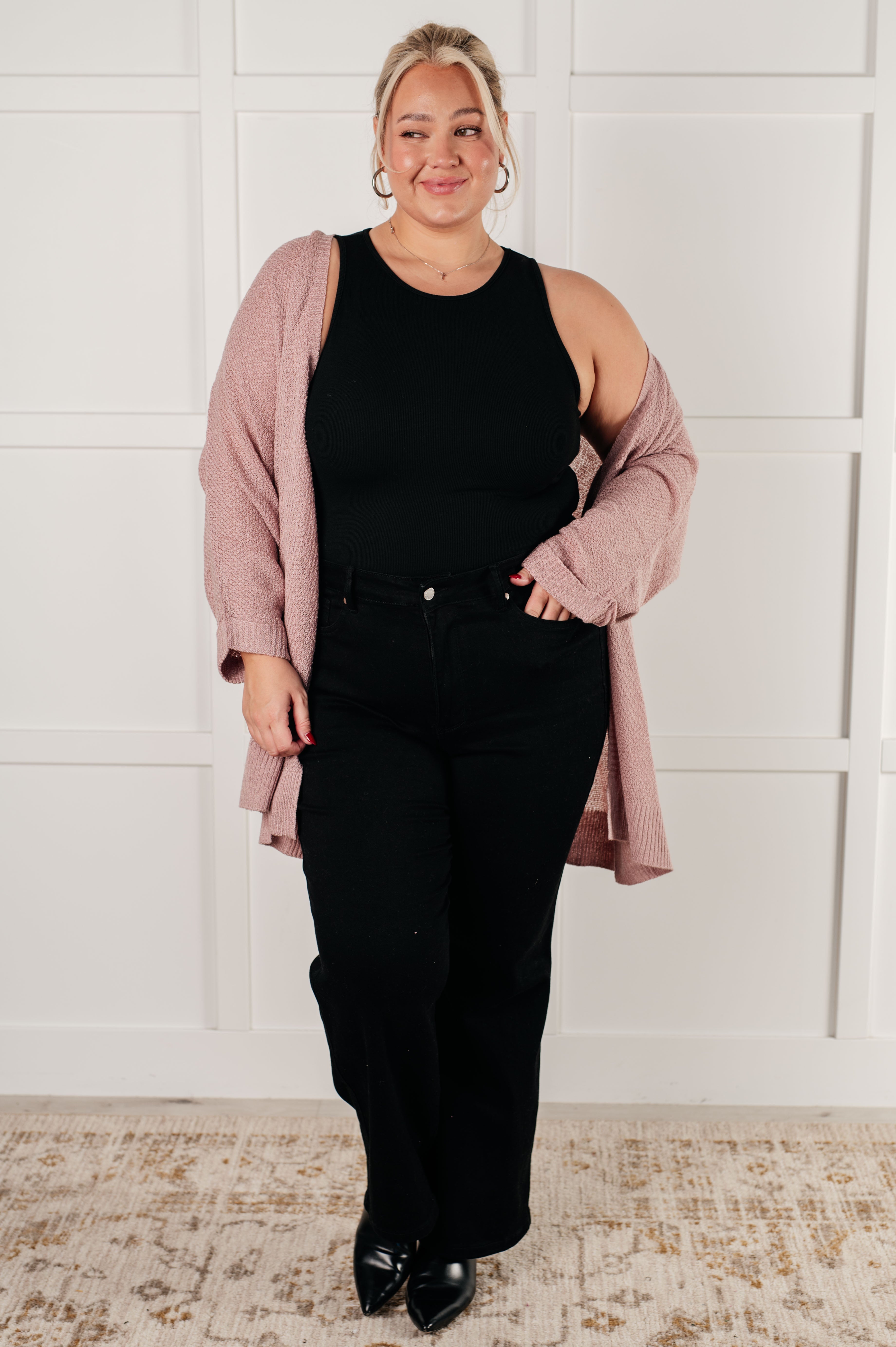 Curvy woman wearing a soft pink open-front cardigan over a sleeveless black top and high-waisted black pants, standing indoors.