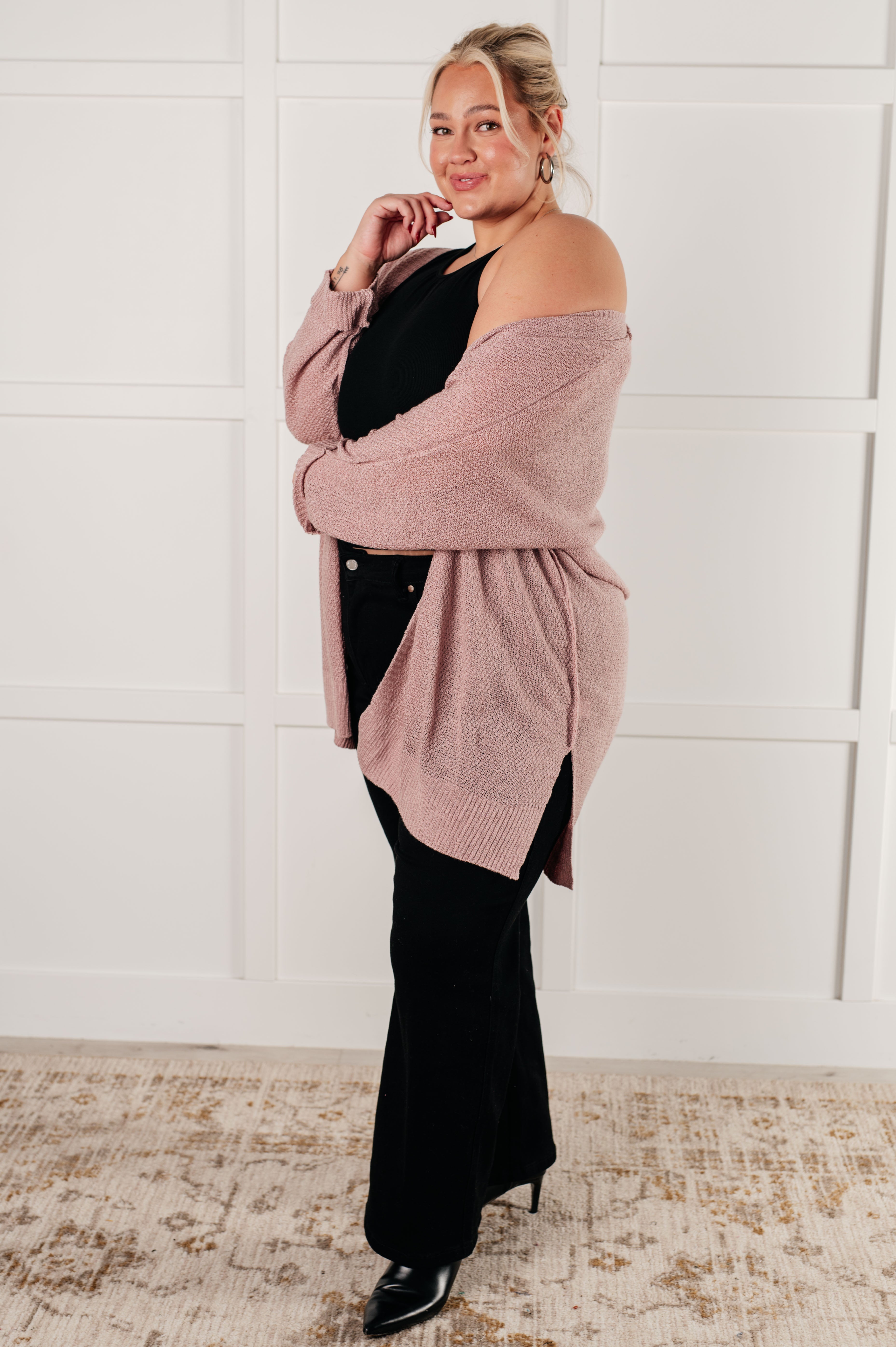 Curvy woman wearing a soft pink cardigan draped off one shoulder over a black top and black pants, standing indoors on a patterned rug.