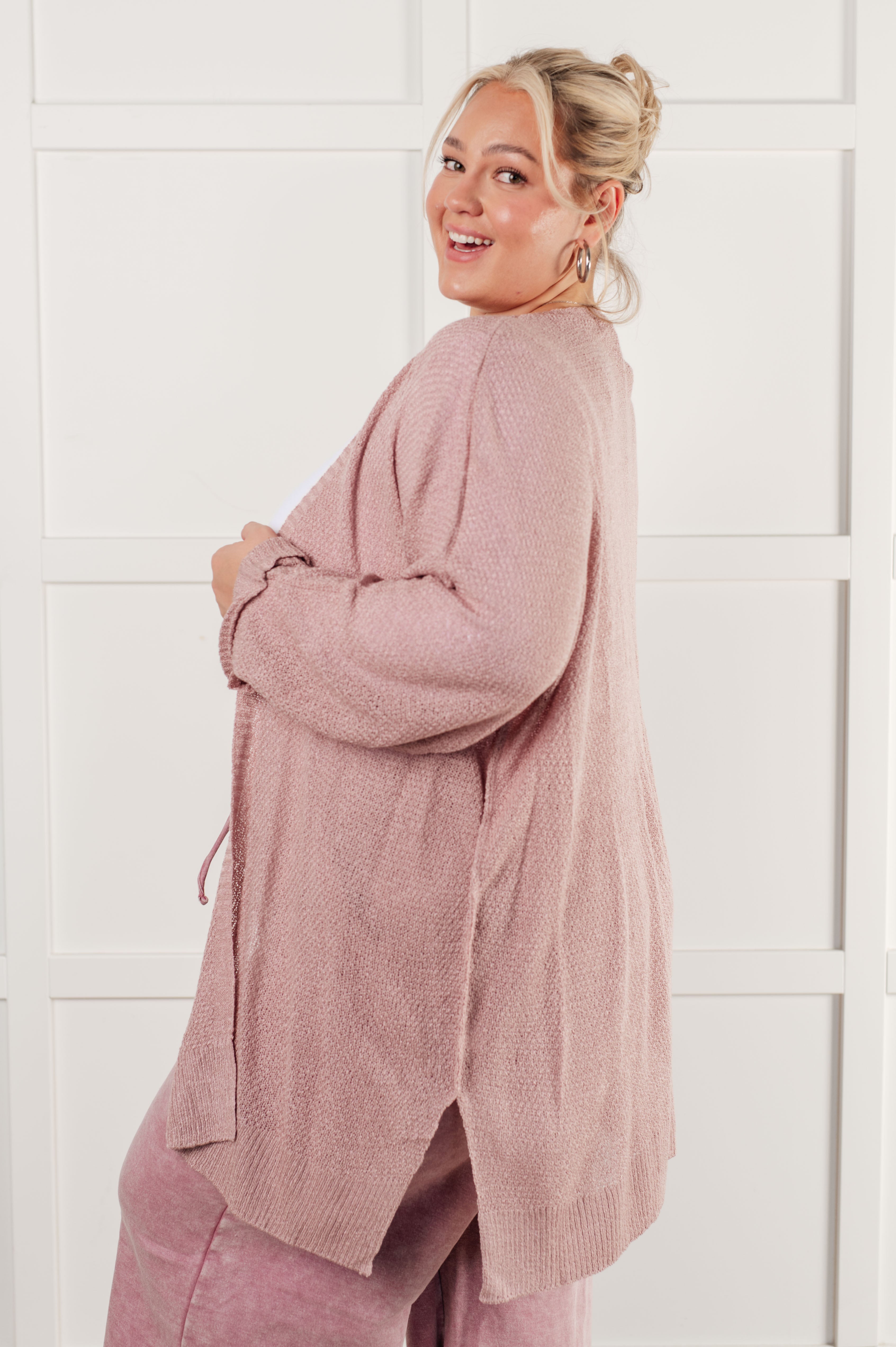 Smiling woman wearing a cozy pink open-front cardigan layered over casual pink pants, standing indoors against a white paneled wall.