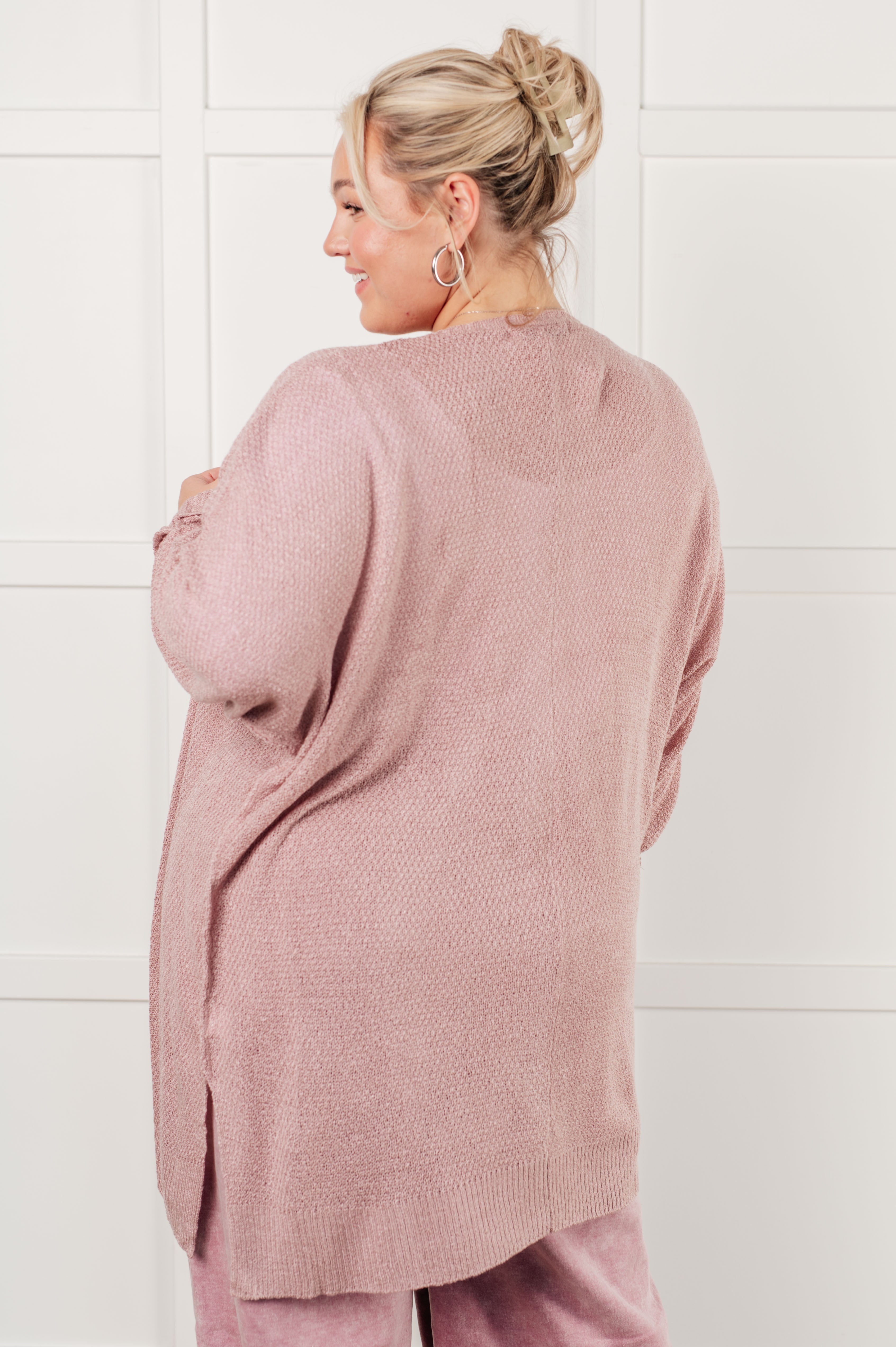 Woman wearing a loose pink open-front cardigan paired with matching pink pants, standing indoors against a white paneled background.