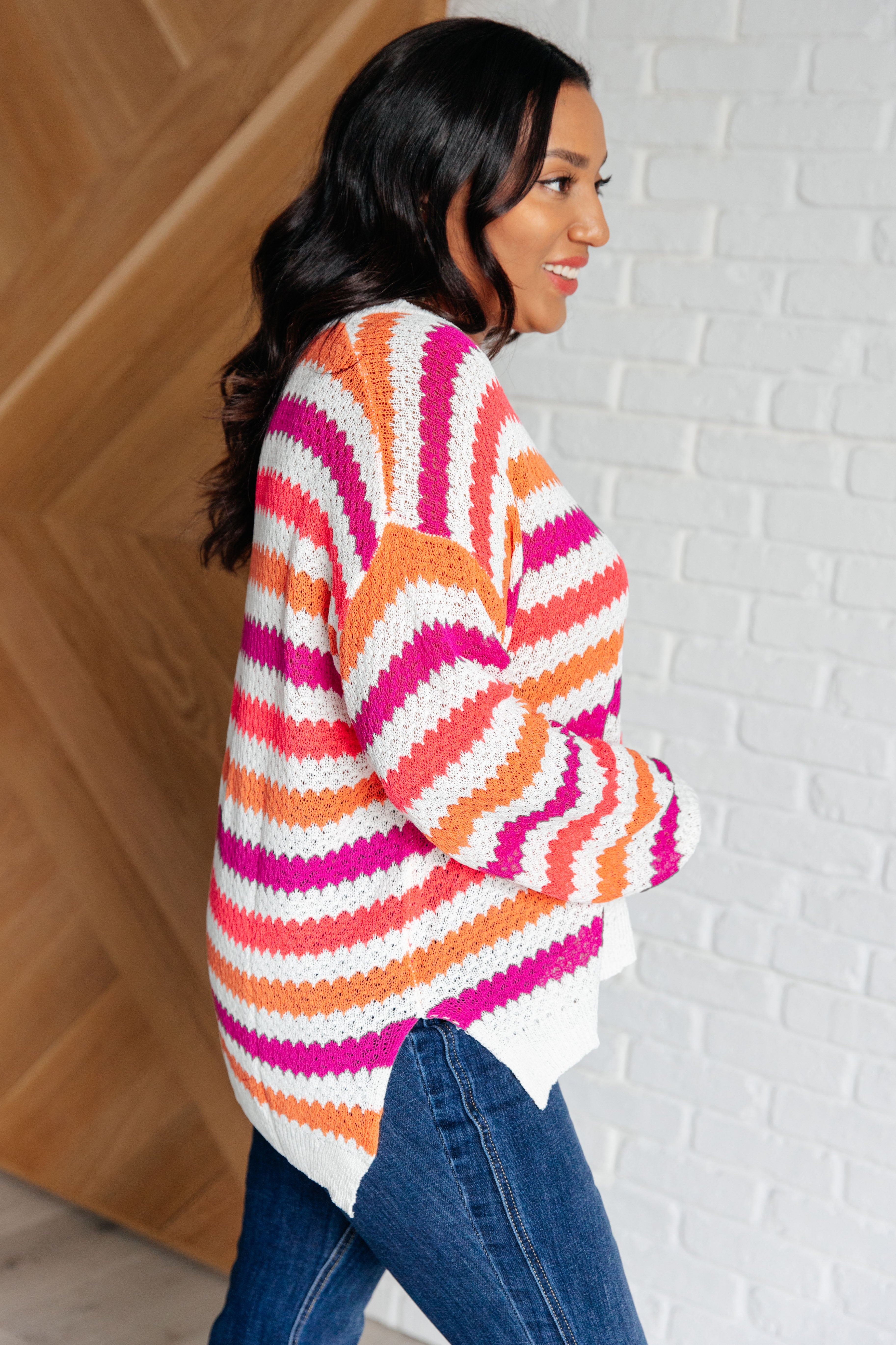 Woman wearing a colorful striped drop shoulder sweater with orange, pink, and white tones paired with dark blue jeans, standing indoors.