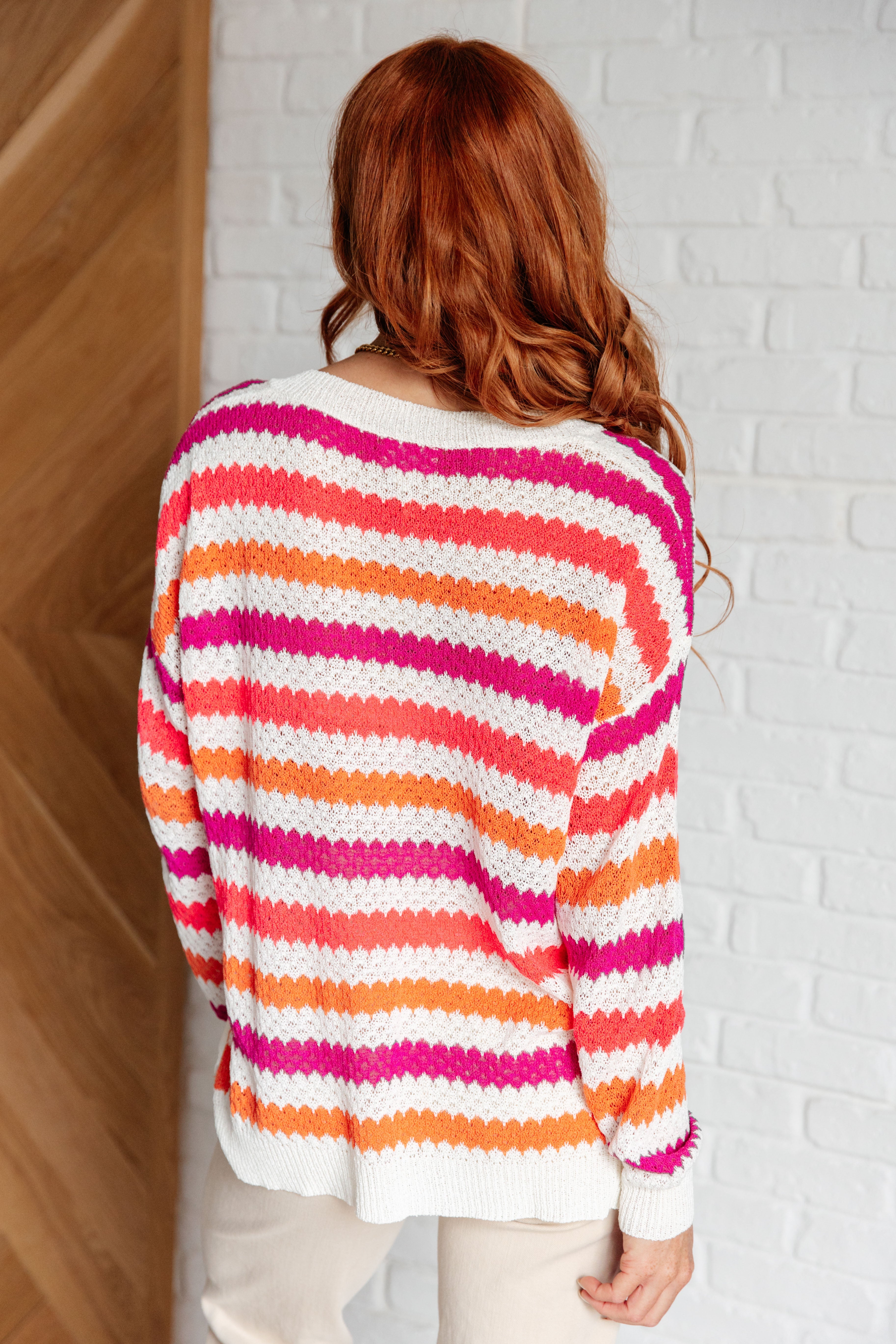 Woman with long red hair seen from behind wearing a knit sweater featuring bright pink, orange, and white stripes paired with light pants indoors.