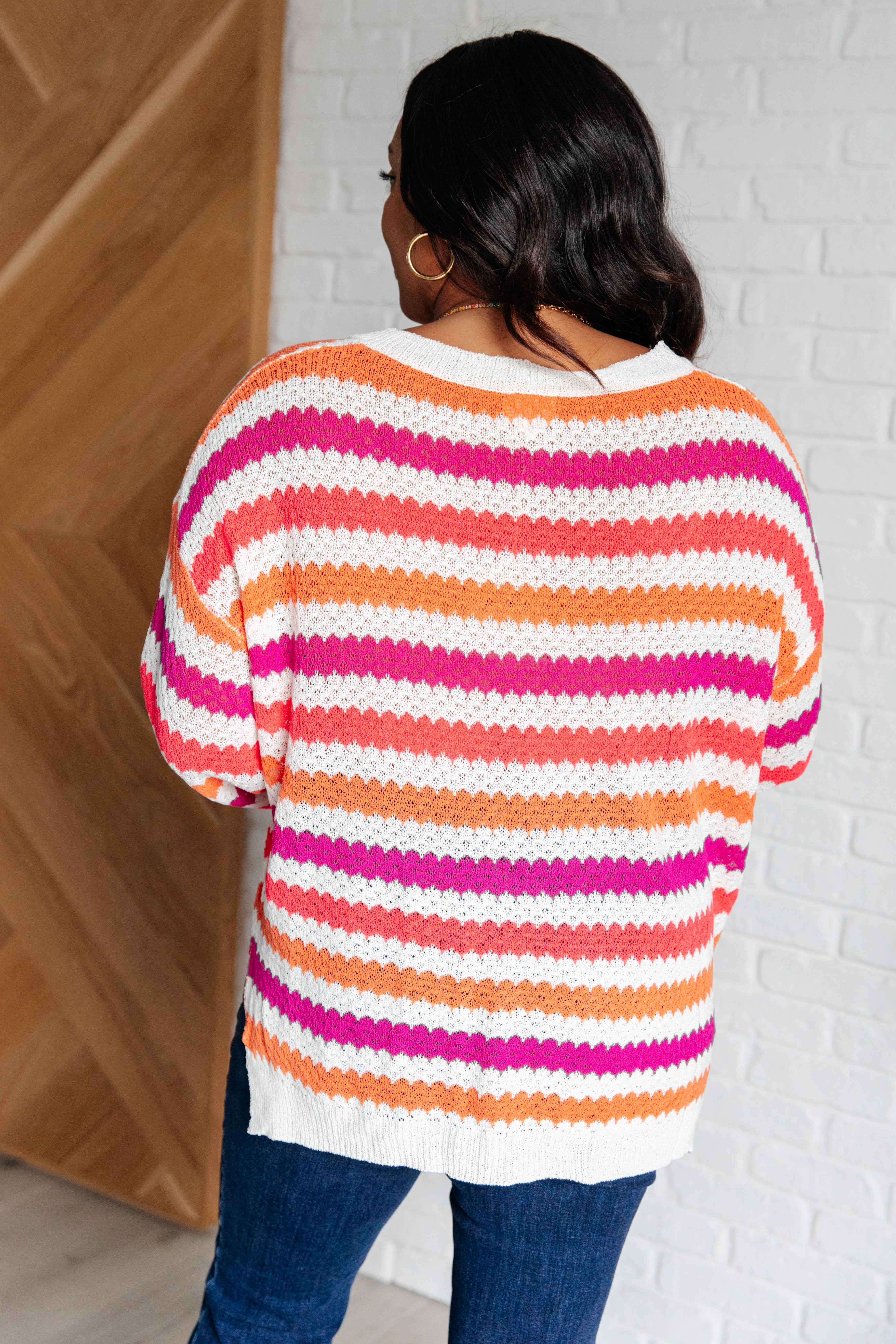 Woman seen from behind wearing a knit drop shoulder sweater with pink, orange, and white stripes paired with dark blue jeans indoors.