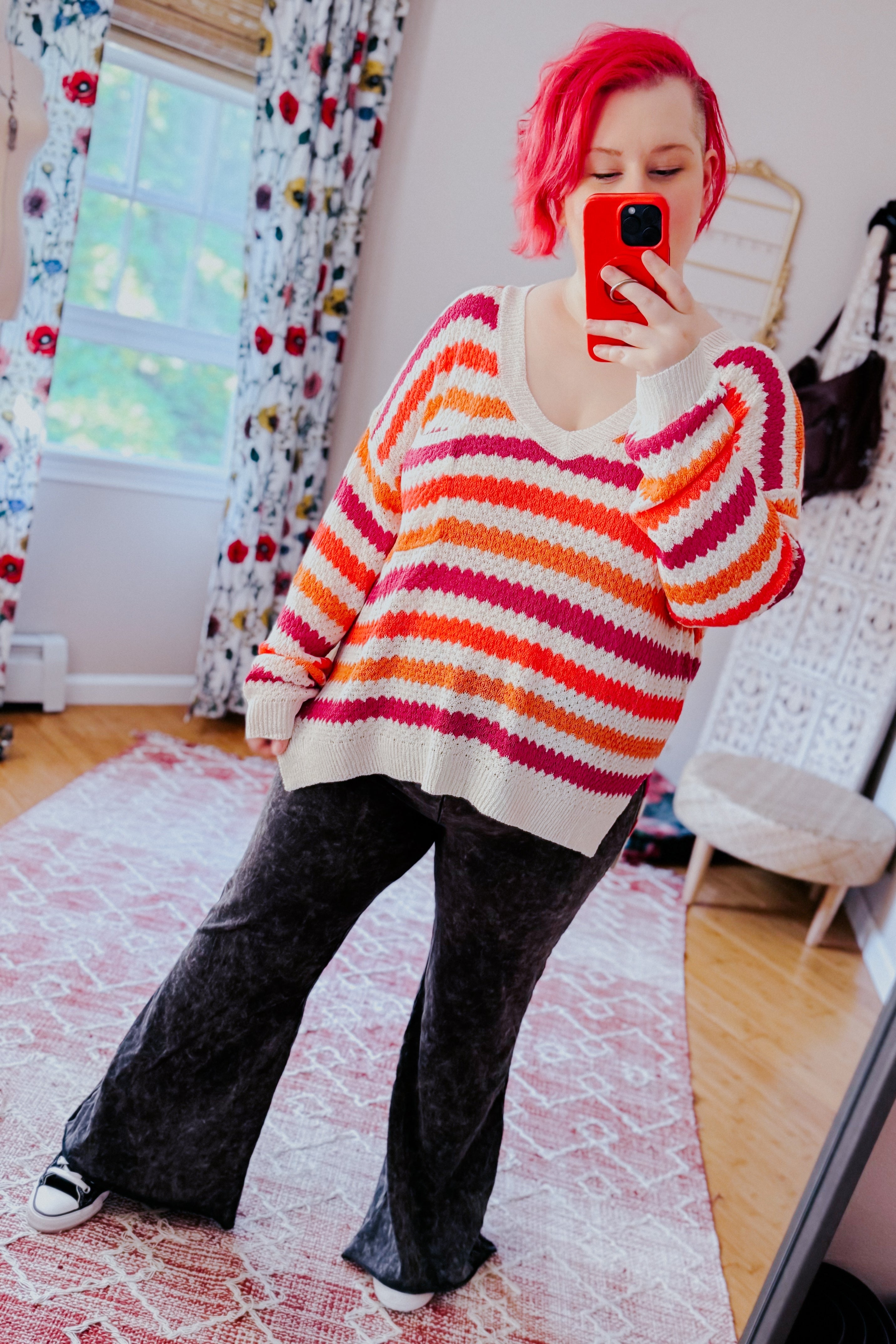 Woman with bright pink hair wearing a pink, orange, and cream striped knit sweater with a relaxed fit paired with loose black pants indoors taking a mirror selfie.