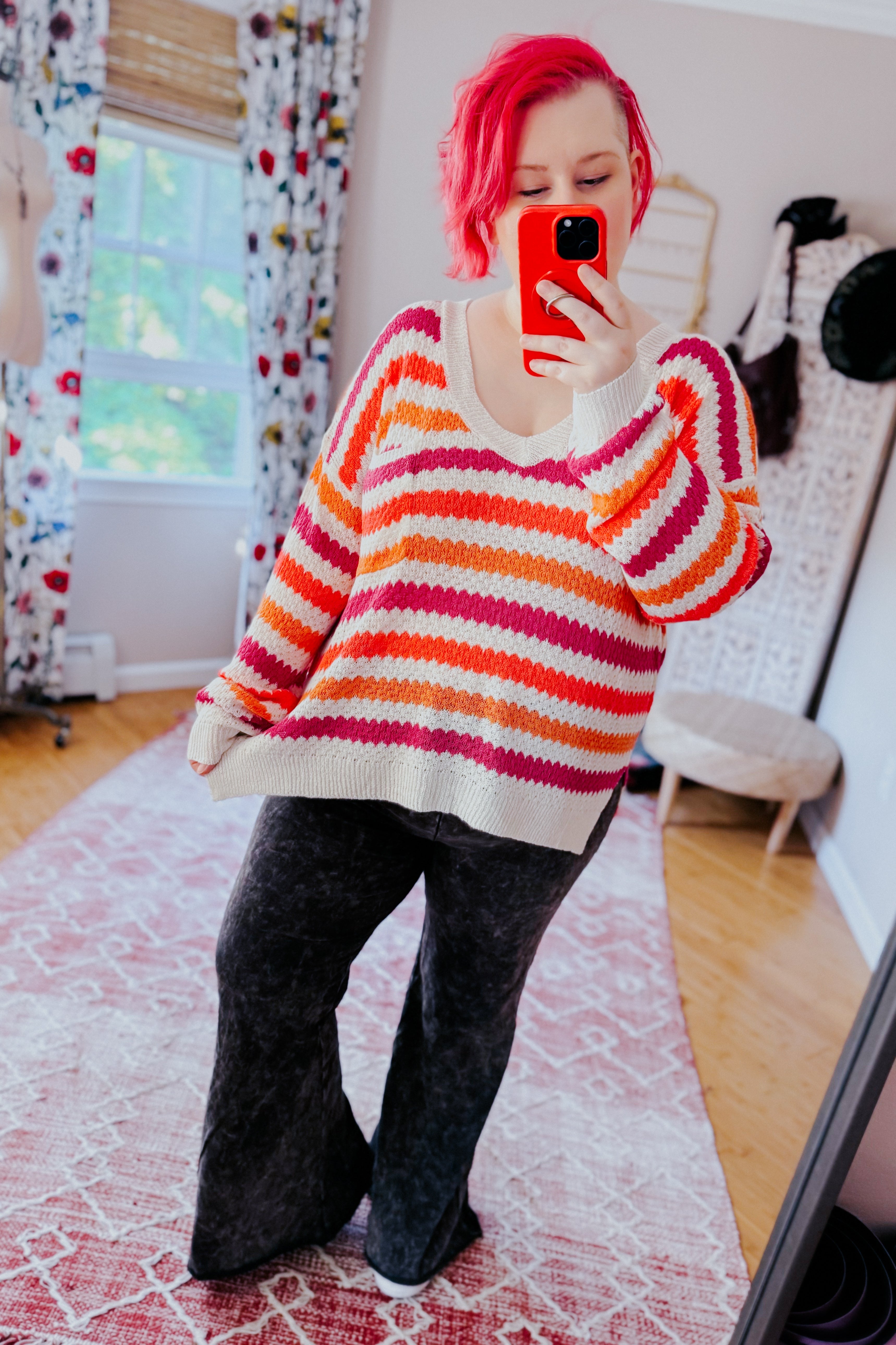Woman wearing a relaxed fit drop shoulder sweater with bold pink and orange stripes paired with wide-leg black pants indoors taking a mirror selfie.