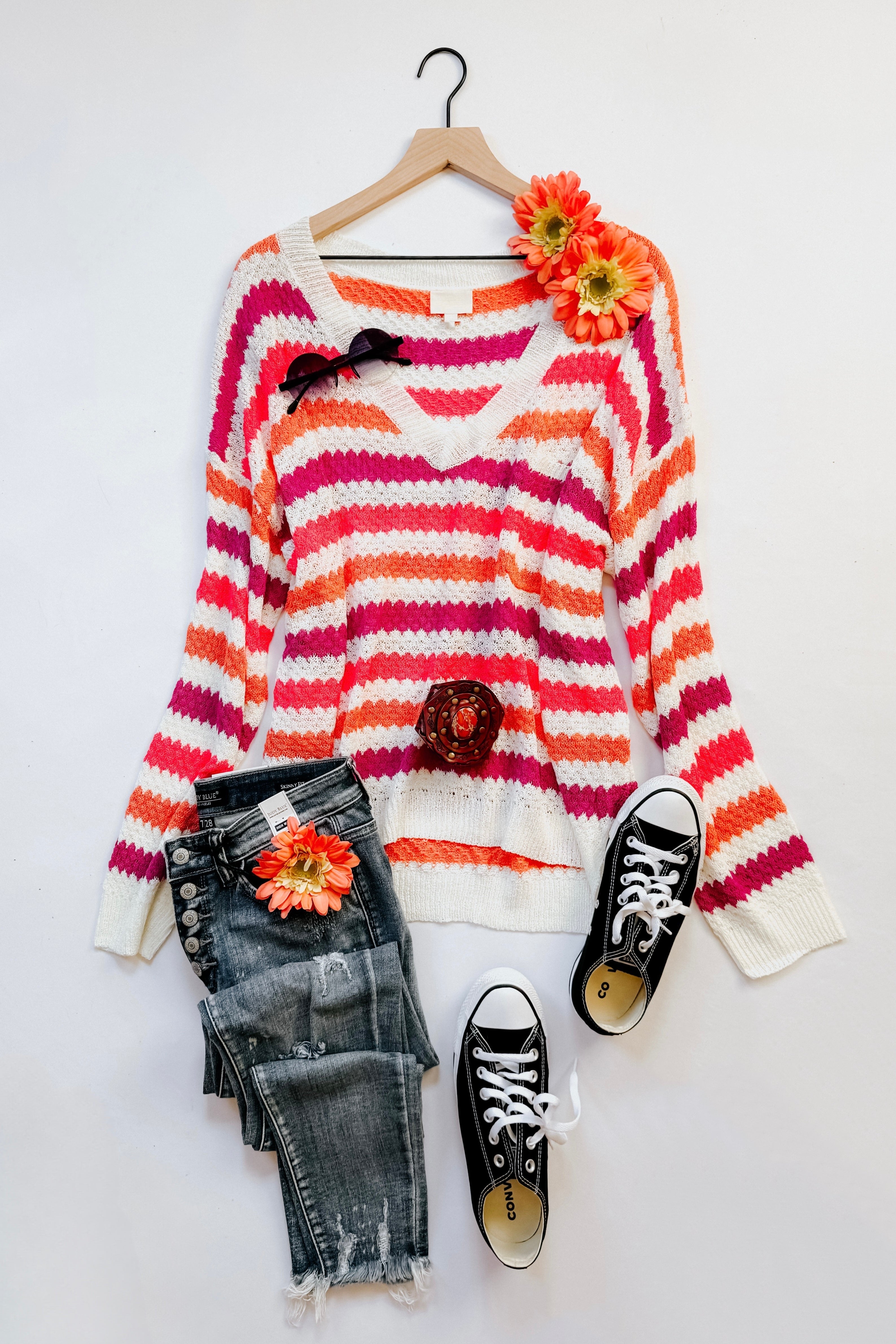 Pink, orange, and white striped knit sweater with a V-neck and drop shoulders displayed with distressed dark gray jeans, black sneakers, and floral accessories.