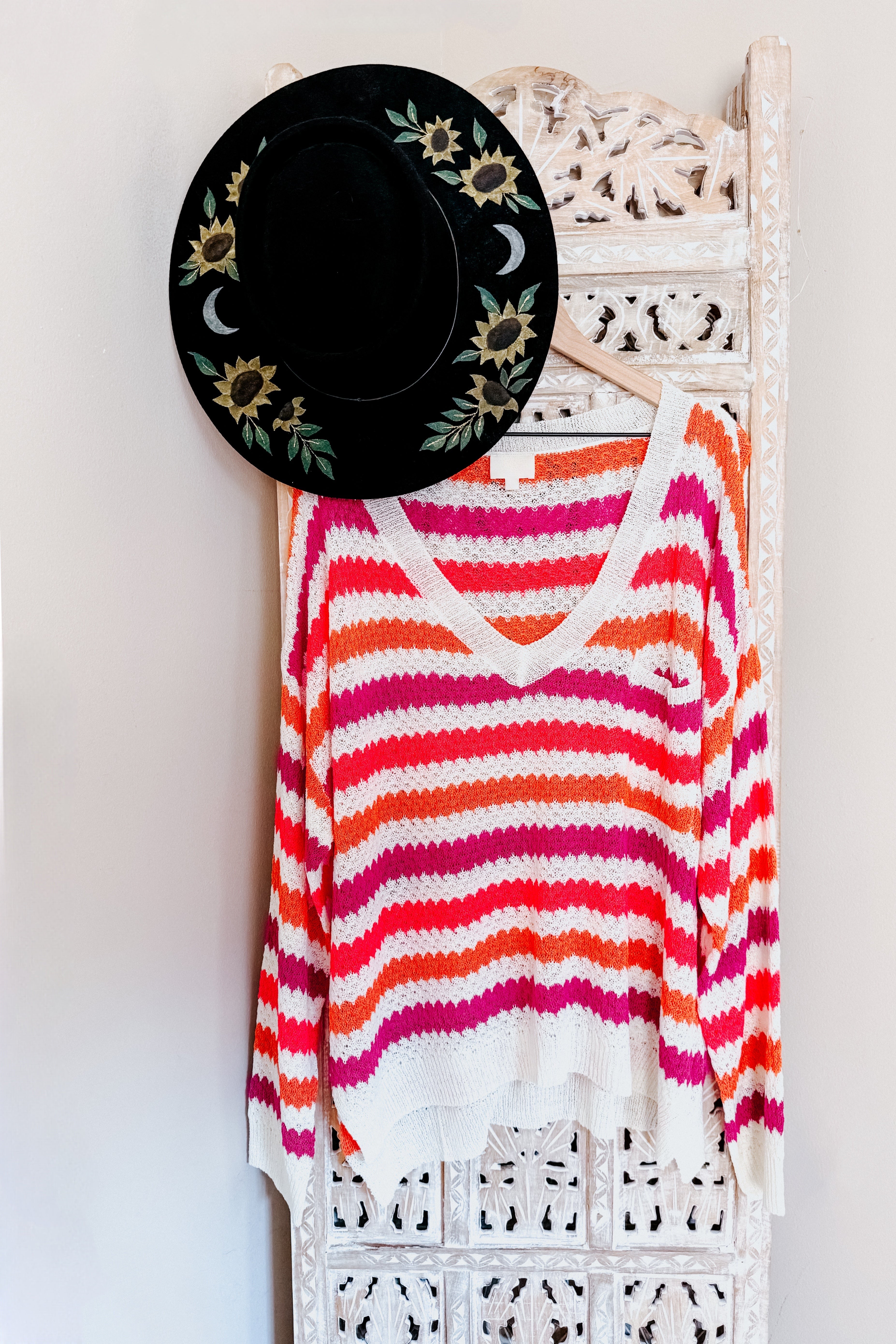 Pink and orange striped knit sweater hanging on a carved wooden screen with a black wide-brim hat decorated with sunflowers and crescent moons.