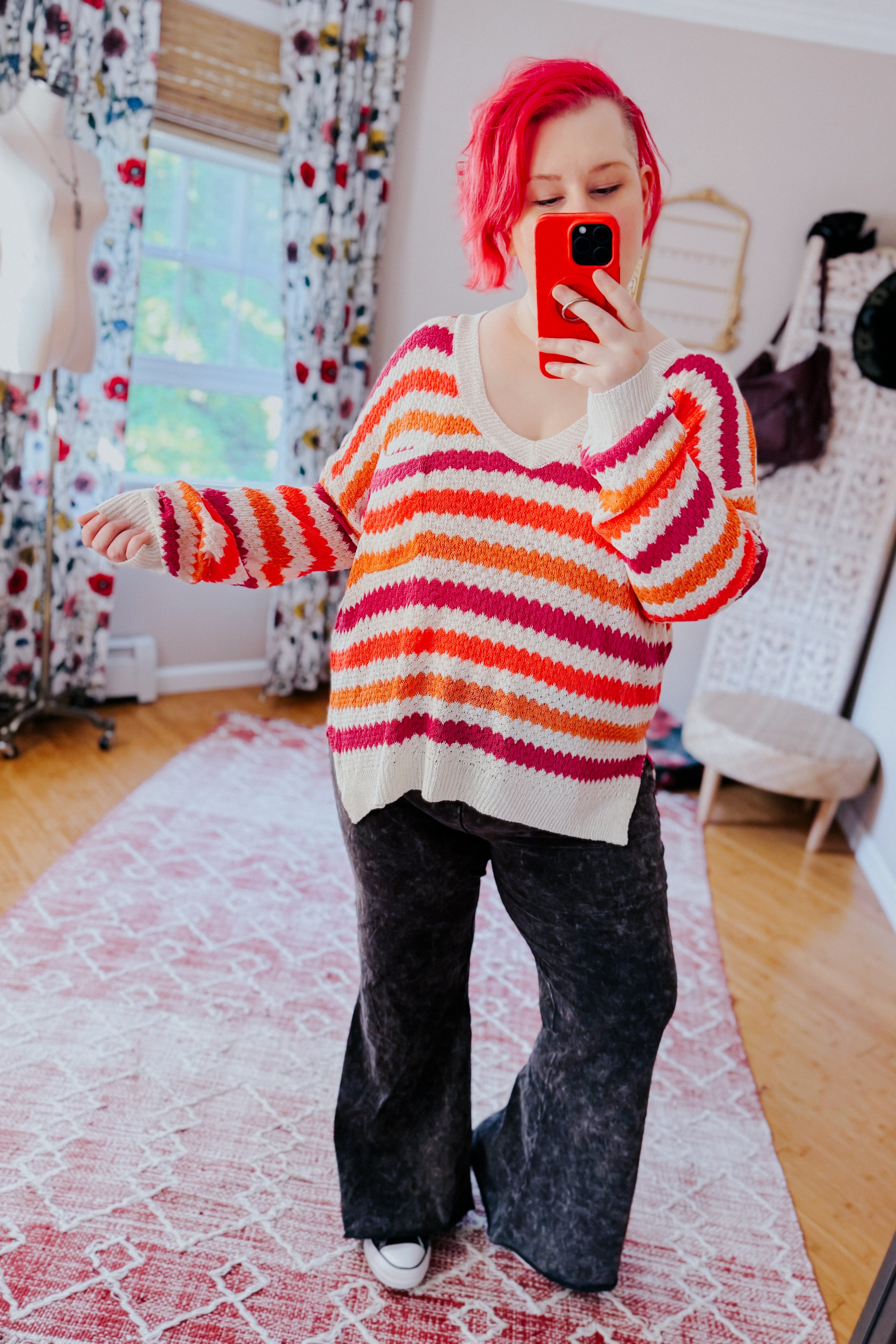 Woman taking a mirror selfie wearing a loose drop shoulder sweater with pink, orange, and white horizontal stripes paired with black wide-leg pants indoors.