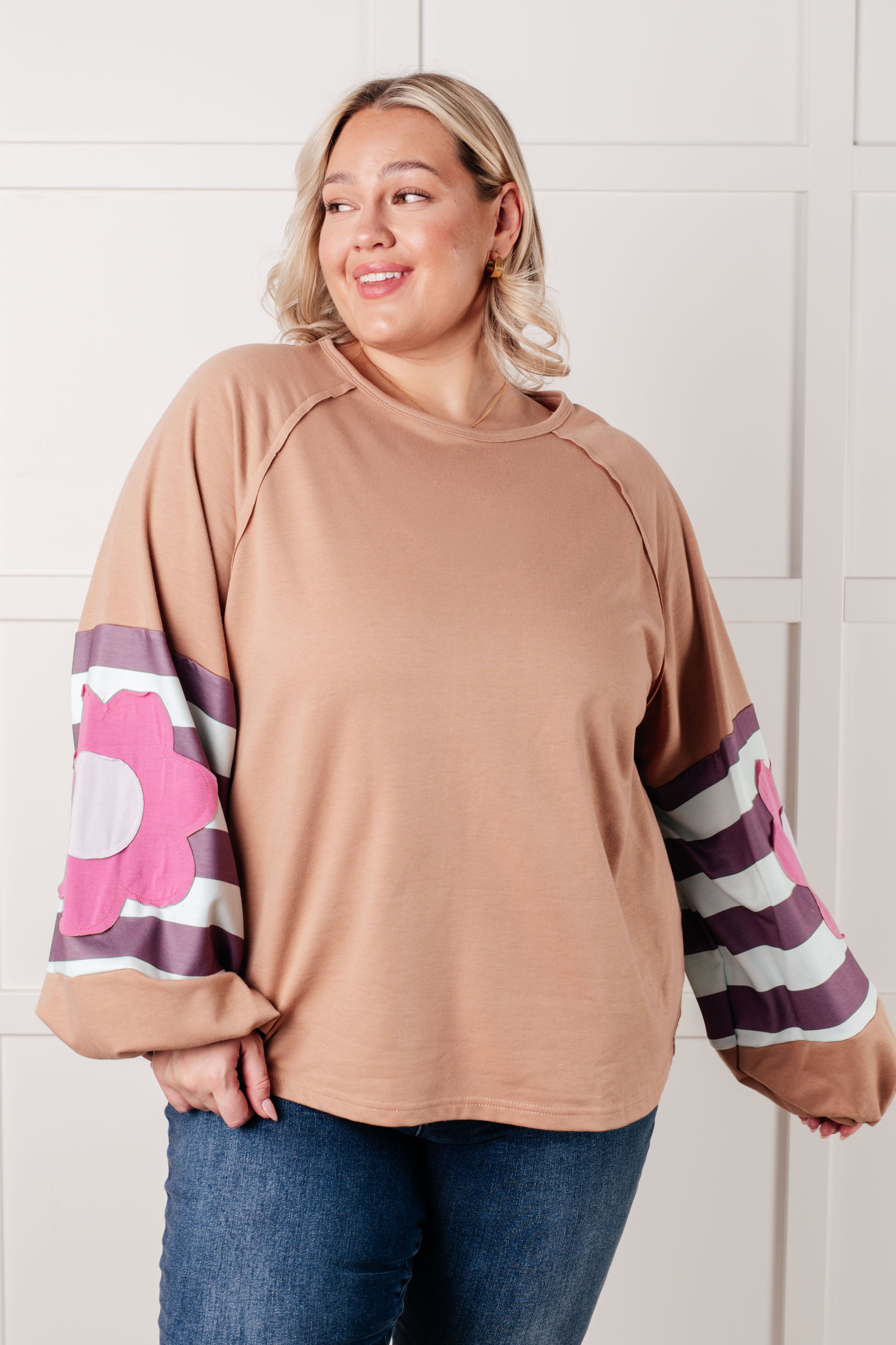 Plus size woman wearing tan top with striped balloon sleeves and large pink floral patch paired with blue jeans indoors