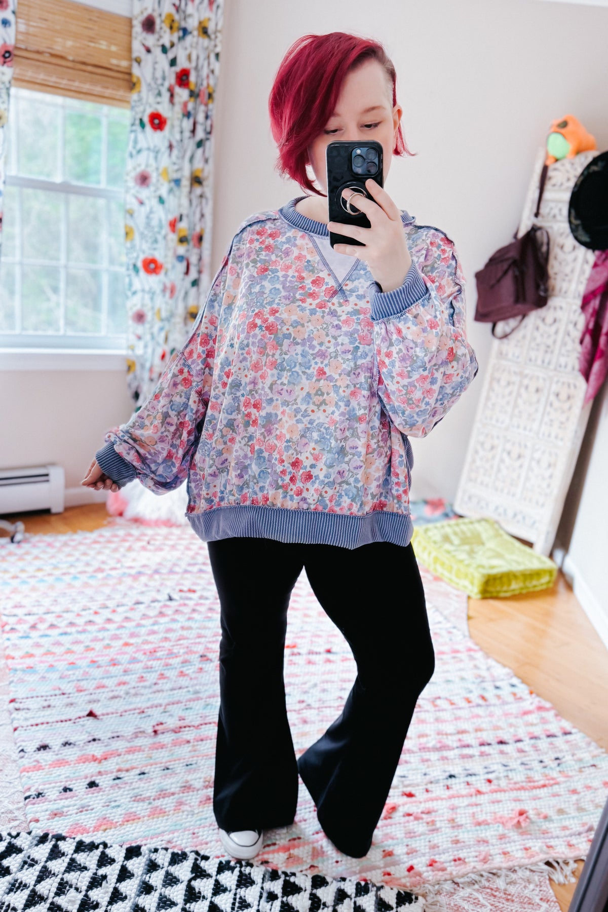 Woman taking a mirror selfie in a blue floral drop shoulder pullover with ribbed trim and black pants in a colorful bedroom