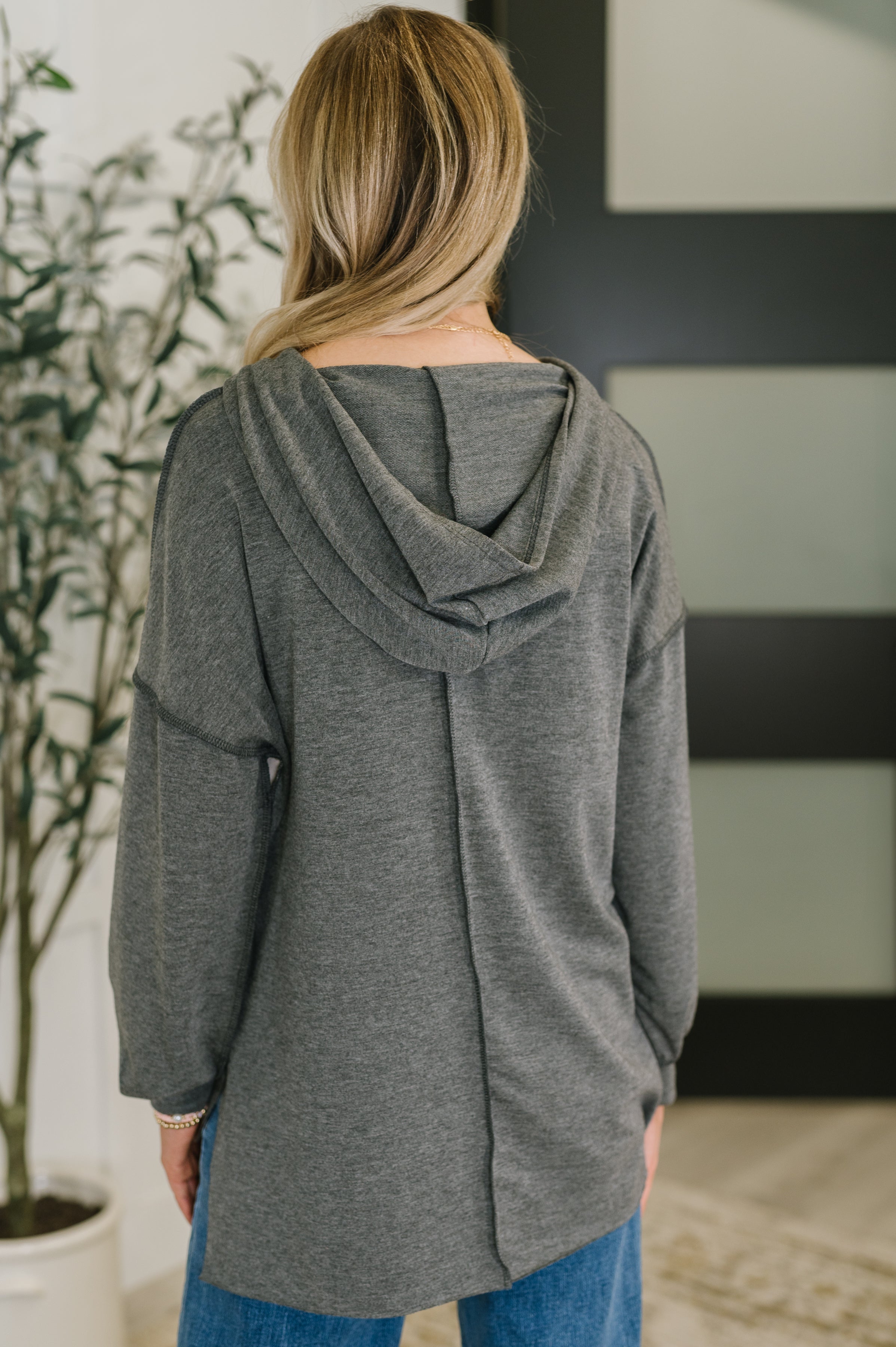 Back view of a woman with blonde hair wearing a loose dark gray hoodie with a relaxed fit and a hood, paired with blue jeans indoors.