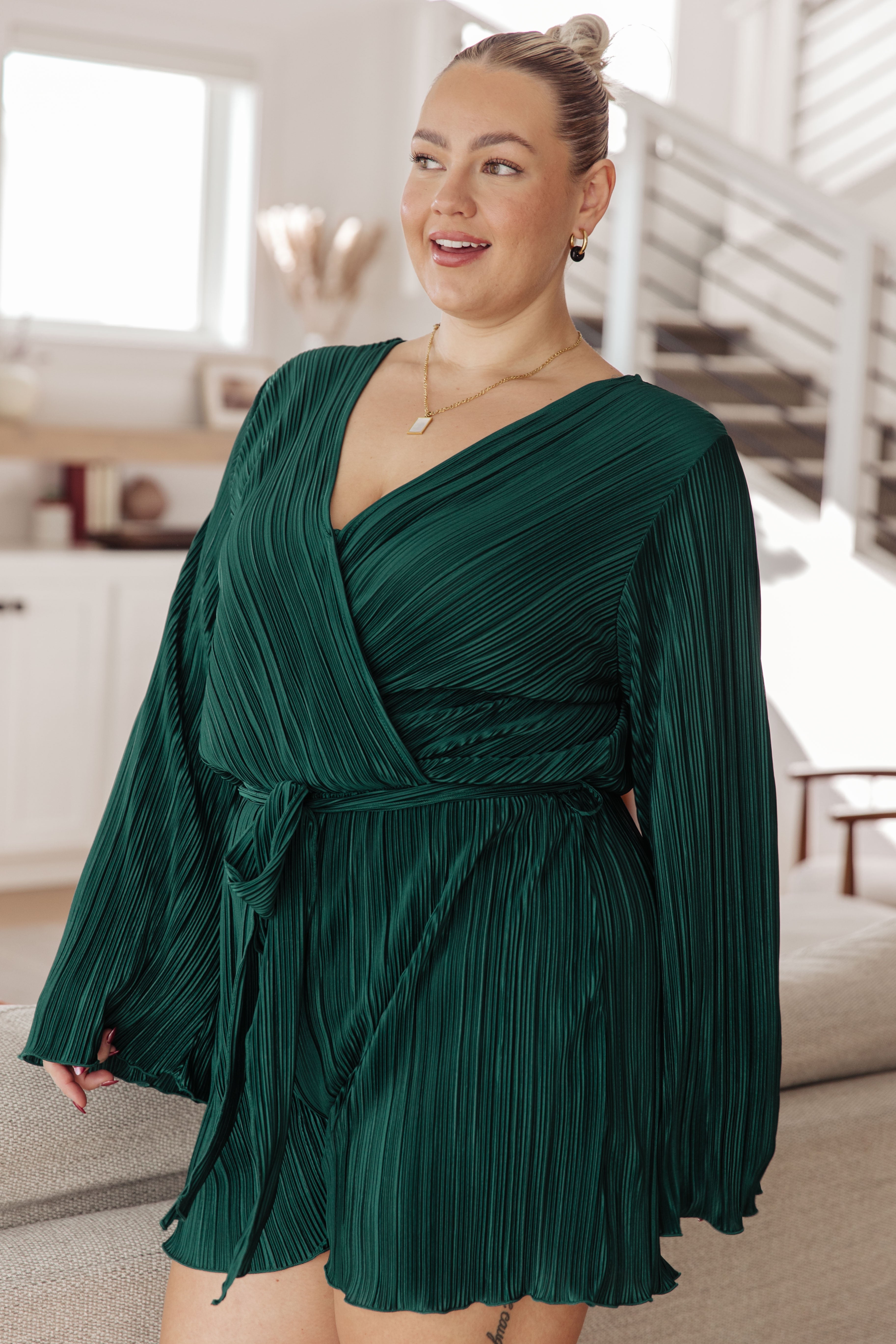 Plus-size woman with blonde hair in a topknot, wearing a hunter green pleated romper with long sleeves and a wrap waist, smiling indoors